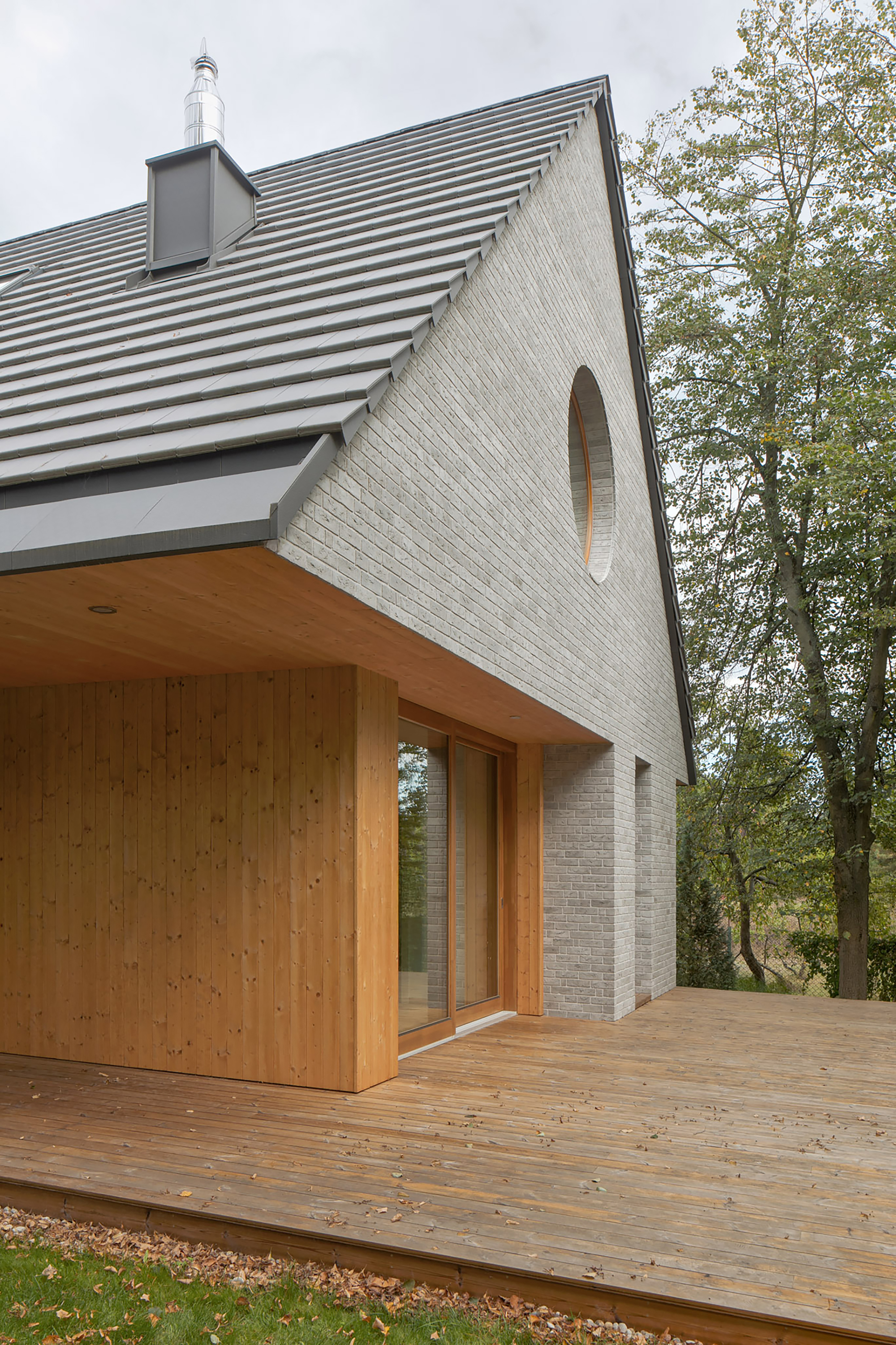 Roof overhang sheltering a timber deck and sliding glass door, with the round gable window and fallen autumn leaves nearby