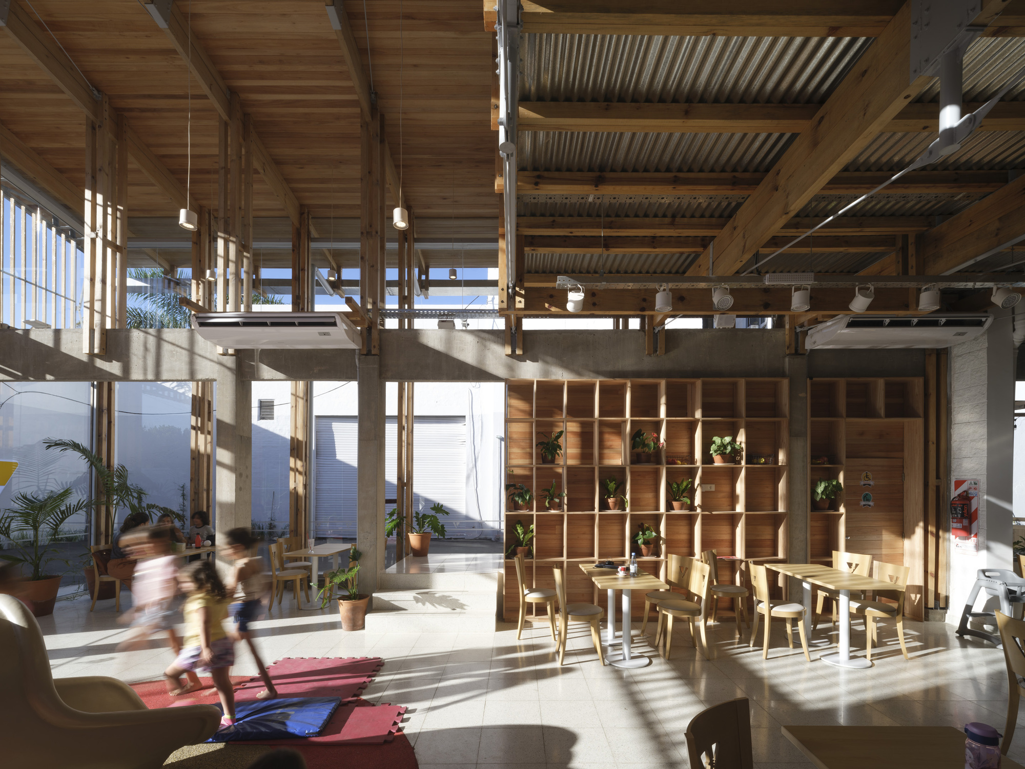 Interior dining hall with timber roof trusses, a plant-filled shelving wall and a blurred child running across a red play mat in afternoon sun