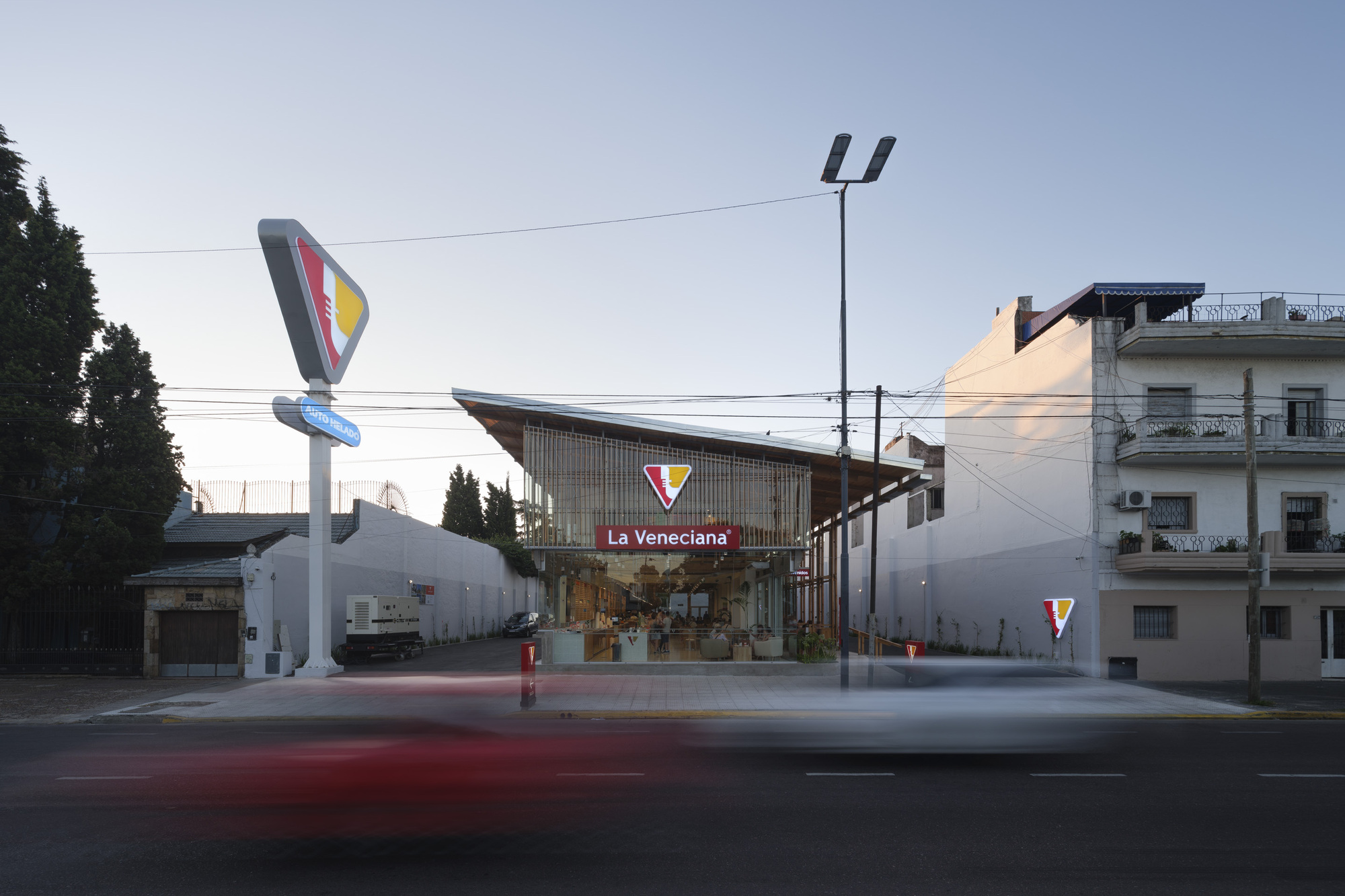 Street view of the glazed corner bakery under a sloping timber roof at dusk, with a tall triangular sign and a blurred car passing