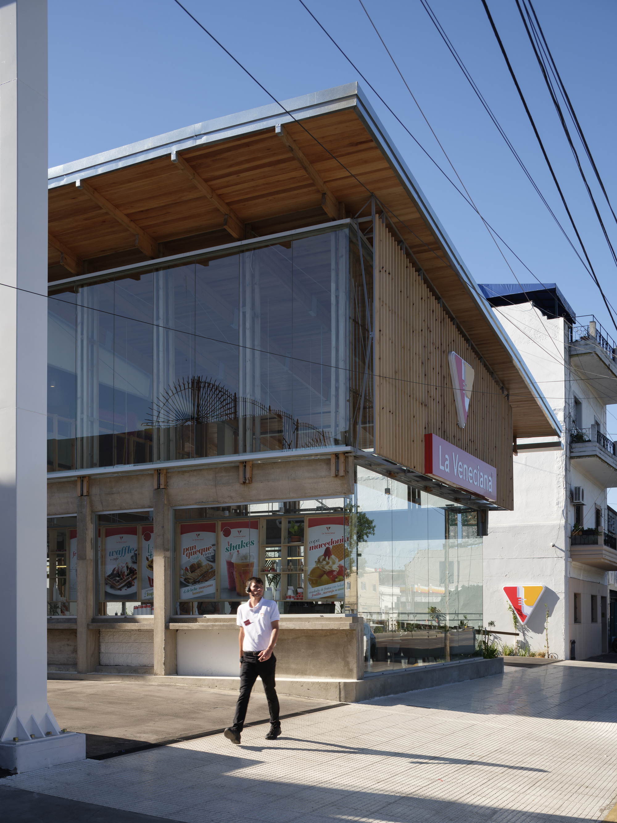 Corner of the two-storey glazed facade with exposed rafters and a timber slat screen, a staff member walking past in bright sun