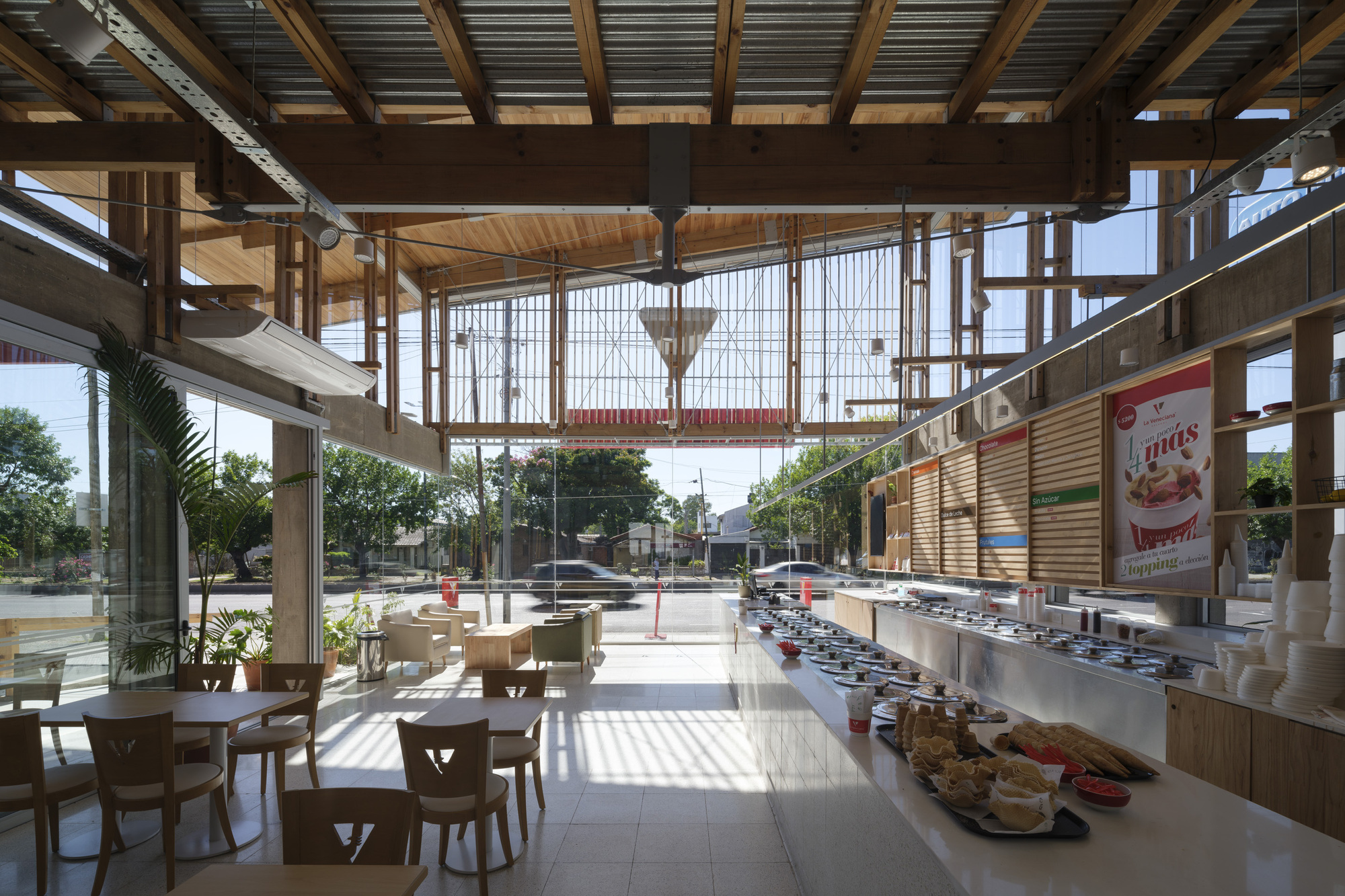 Interior looking out through the glazed front past a food counter and timber tables, with exposed roof beams and street trees beyond