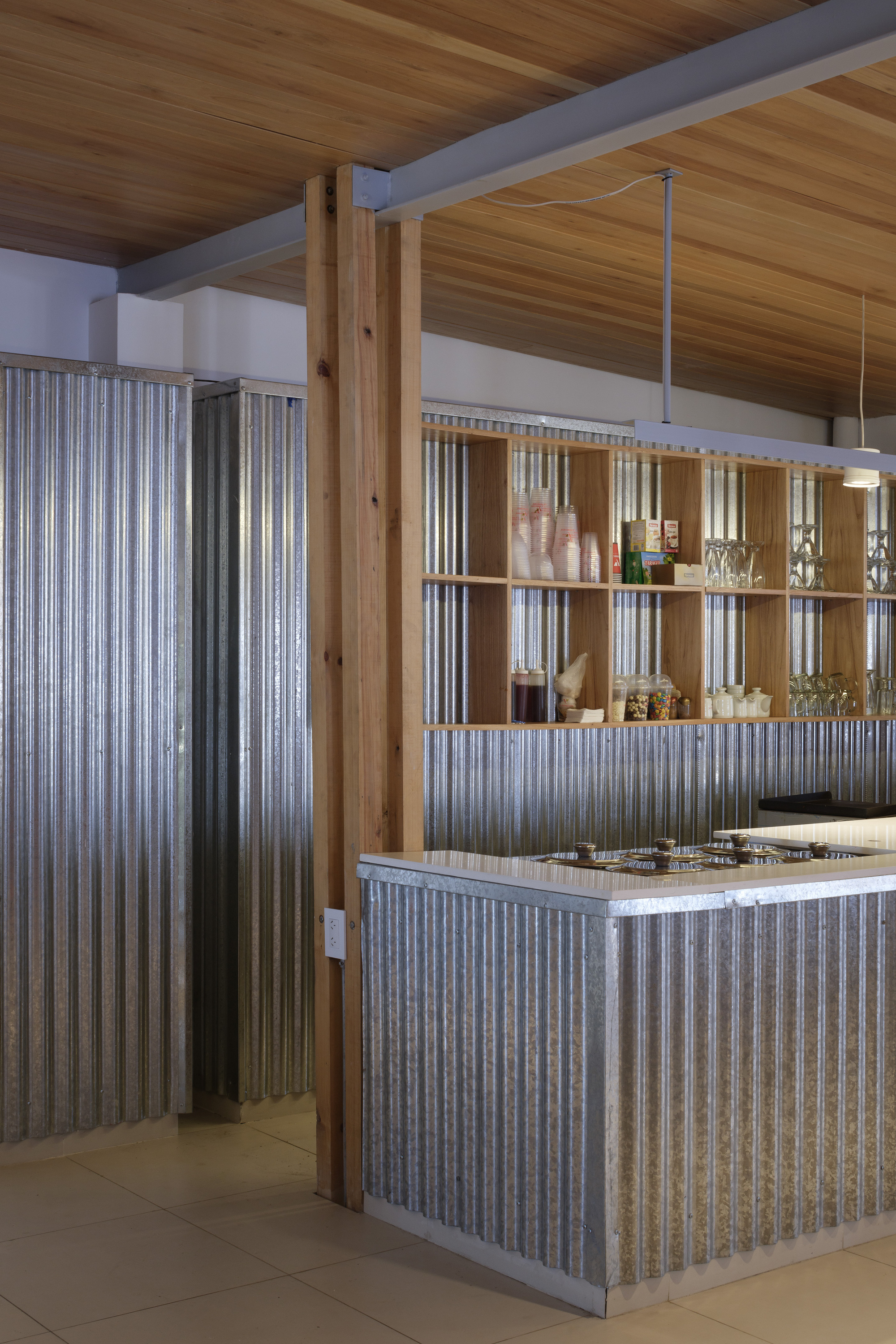Corrugated galvanized steel panels clad the counter and walls beside a timber column, with open shelving holding glasses and jars below a plank ceiling