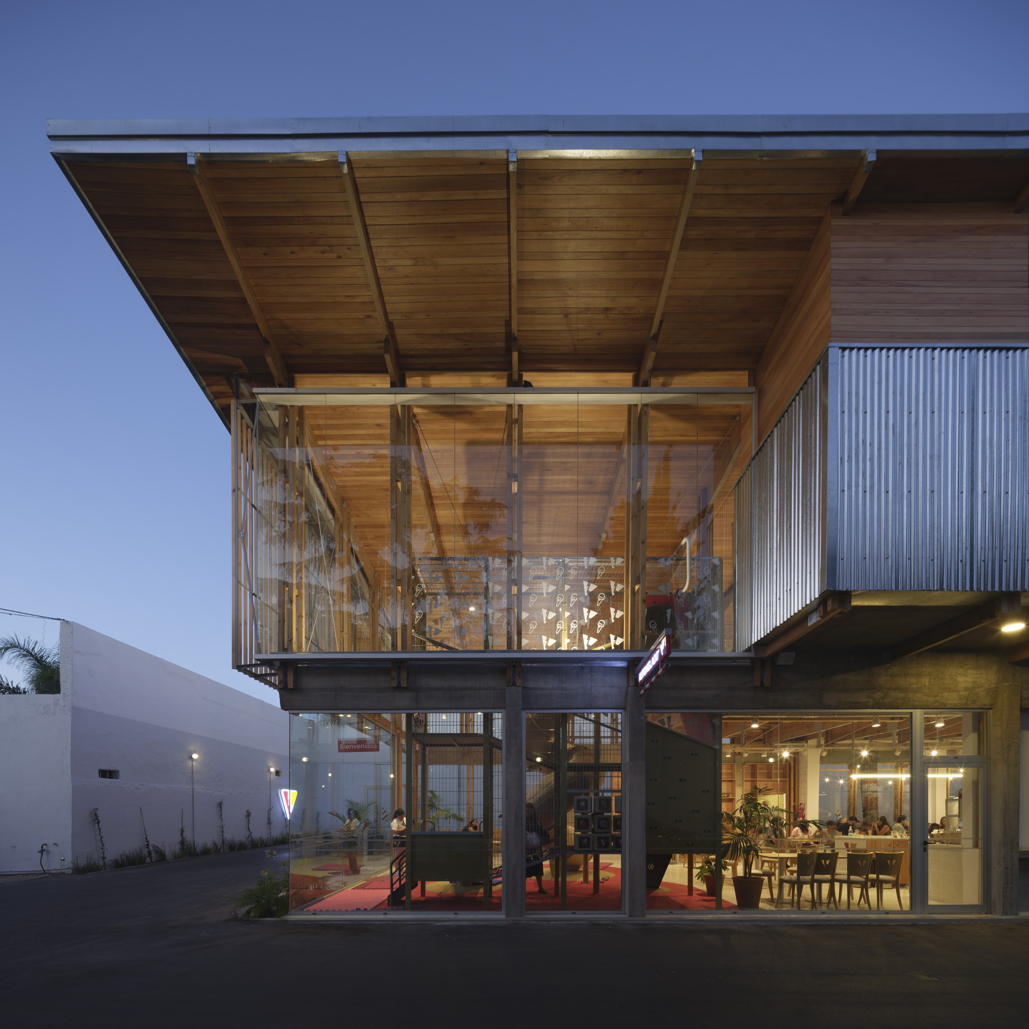 Dusk view of the cantilevered timber roof above a glazed upper floor and a lit ground-level dining room with corrugated metal cladding
