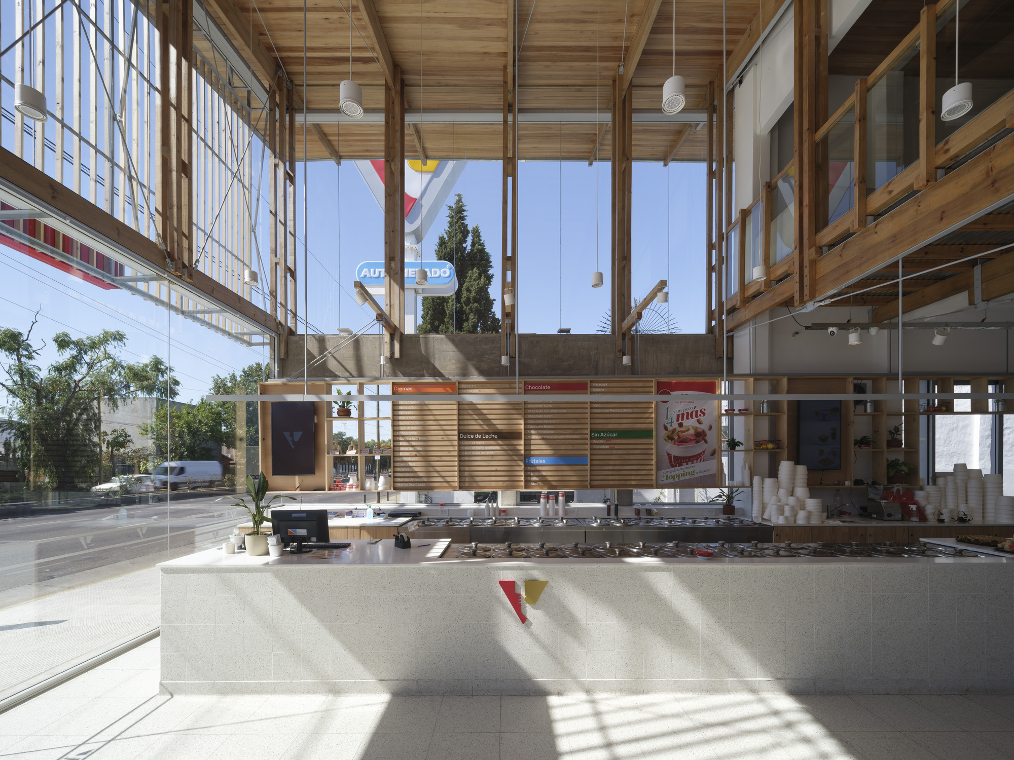 Ice cream counter with terrazzo-clad front and wood slat menu wall, beneath tall timber columns and sunlit glazing
