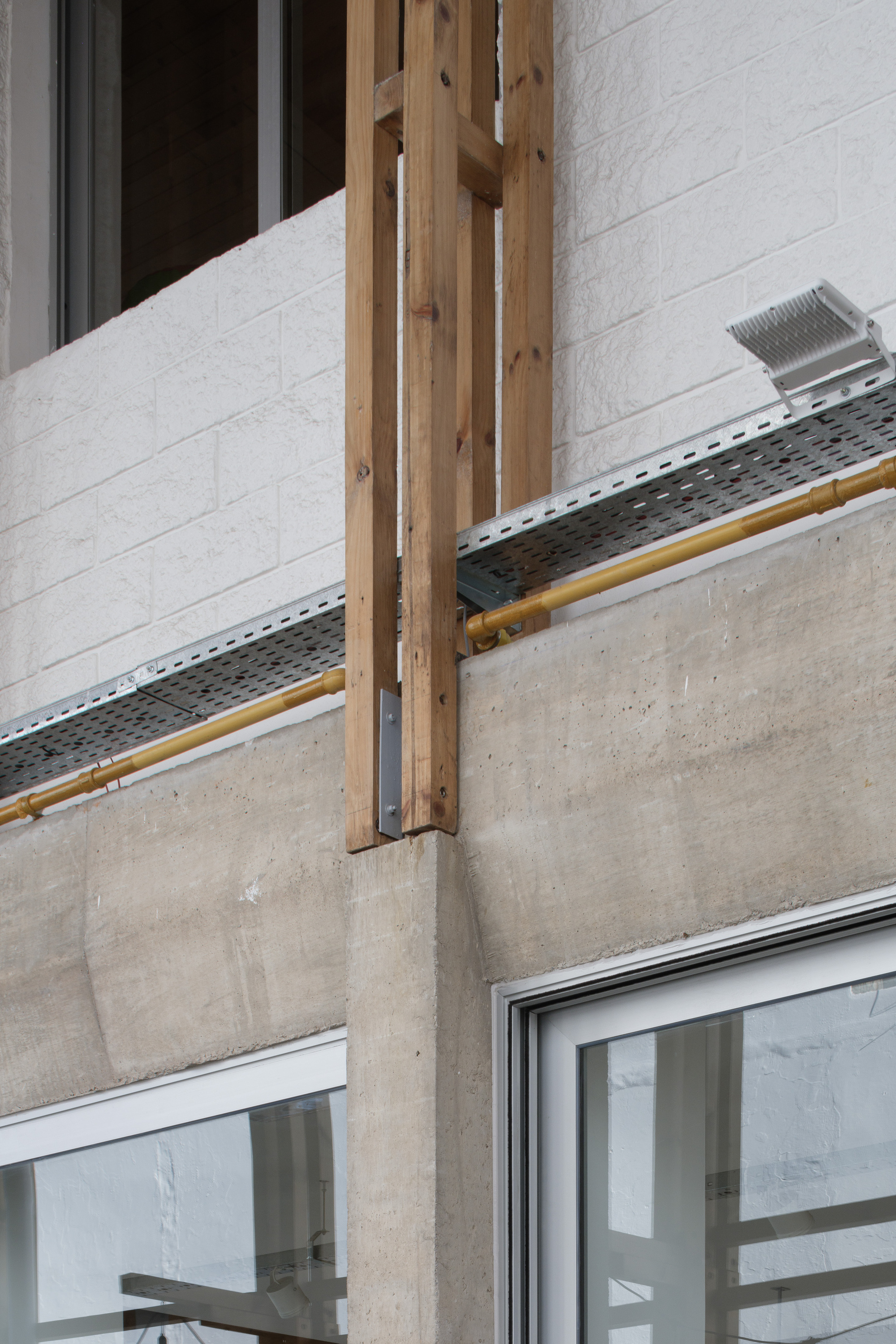 Detail of a wooden post bolted to a concrete beam, with a steel cable tray and yellow pipe running past white brick