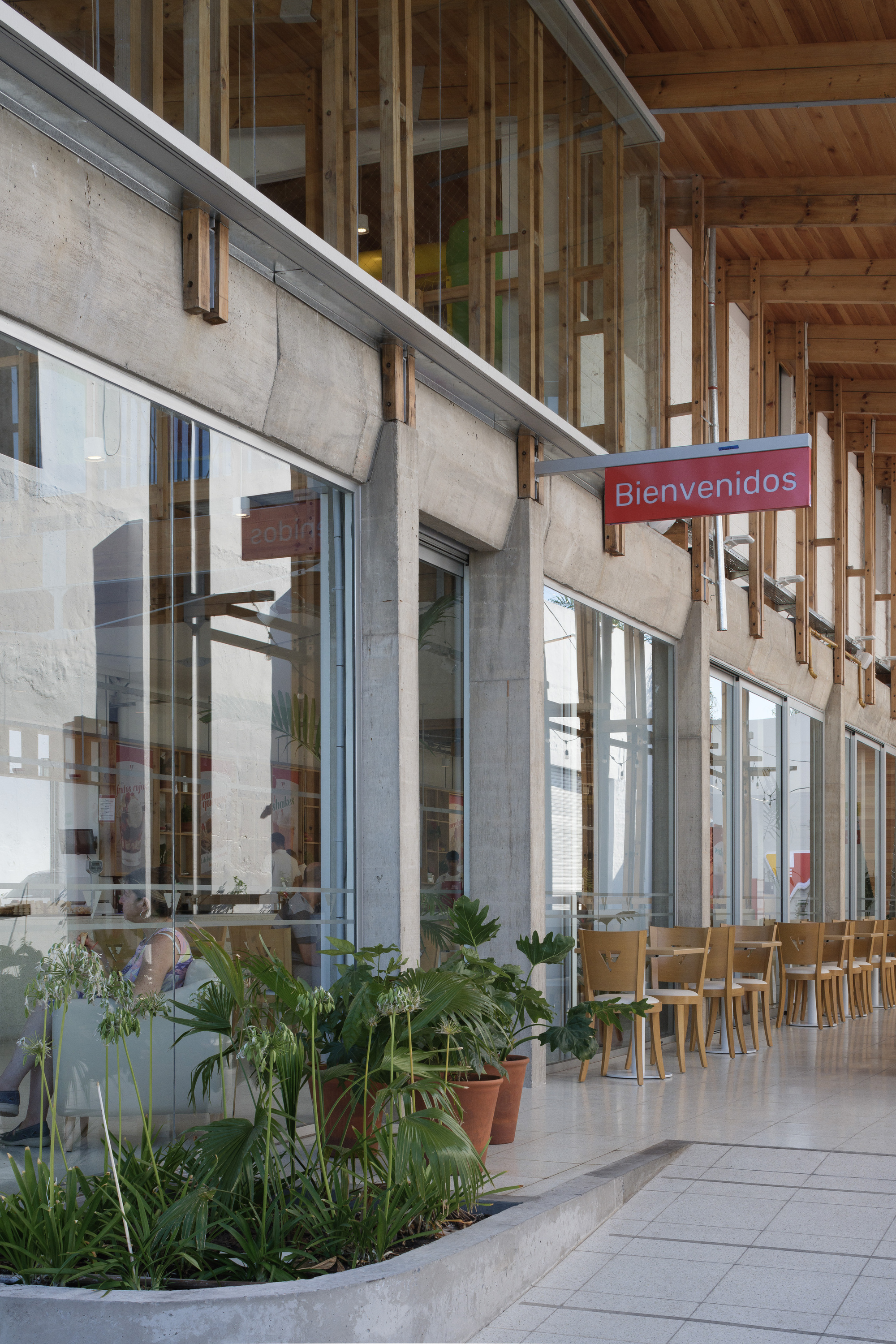 Concrete frame and glazed cafe edge with a Bienvenidos sign, potted plants in a planter and wooden chairs on a tiled floor