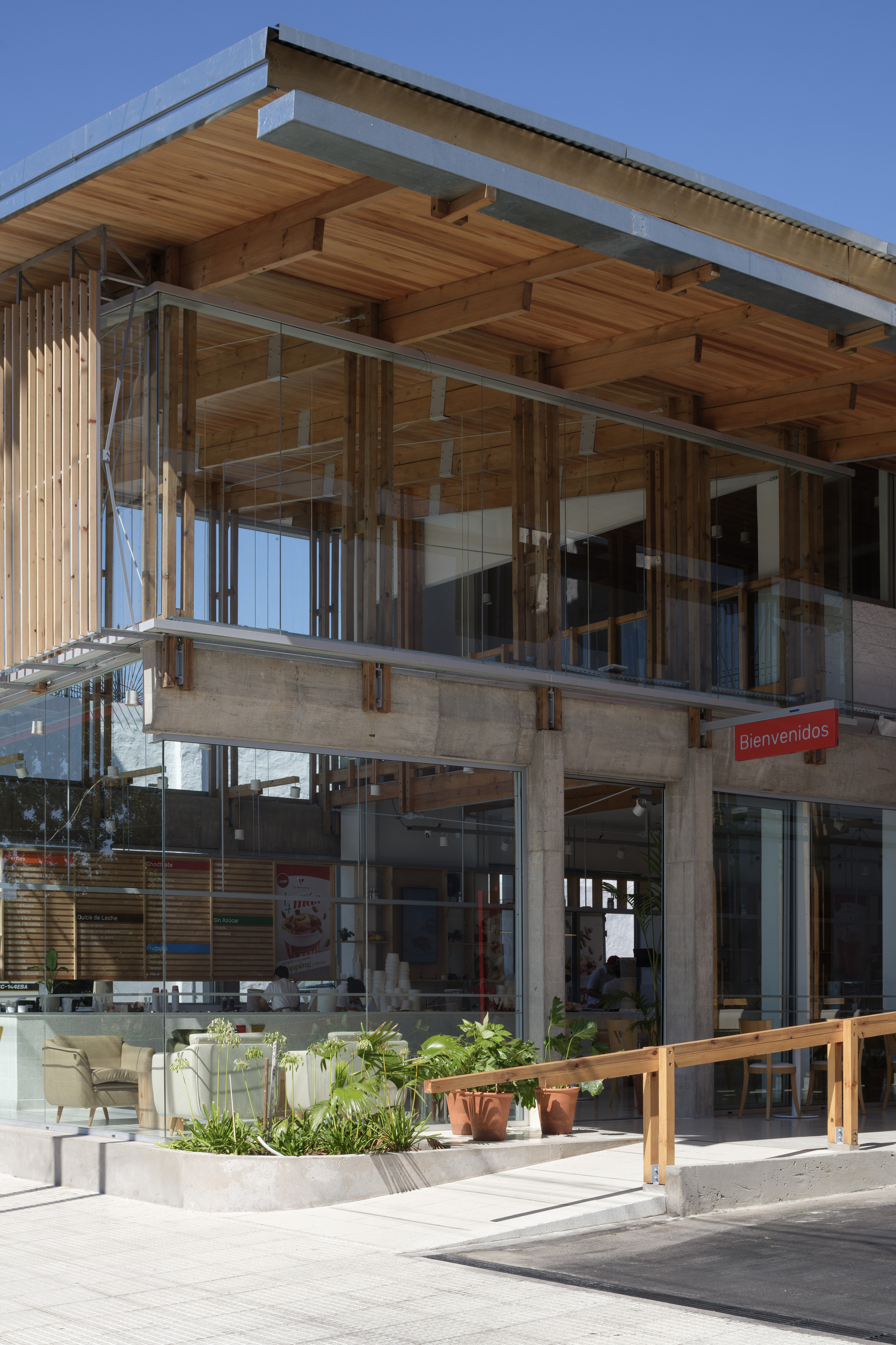 Corner of the two-storey cafe with a timber roof soffit over glass walls, a wooden railing and planted bed under blue sky