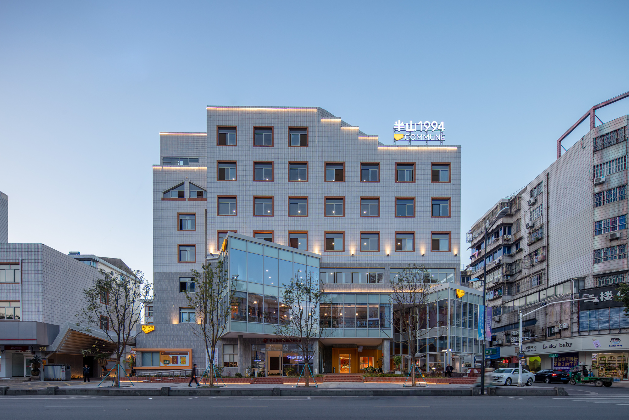 Street facade of the grey tiled block with square red-framed windows and a glass base, lit warmly at dusk