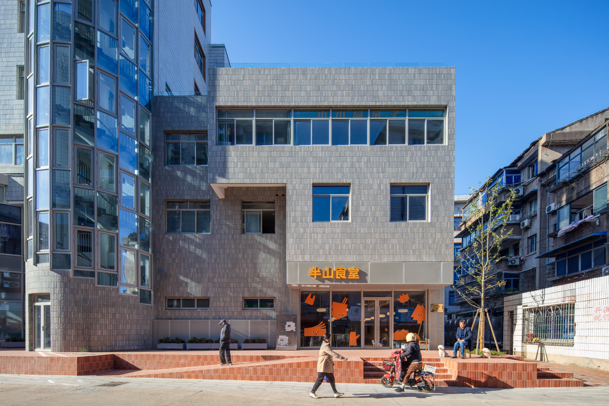 Grey tiled facade with an orange-lettered ground floor entrance above a stepped brick plinth, a person walking past in bright sun