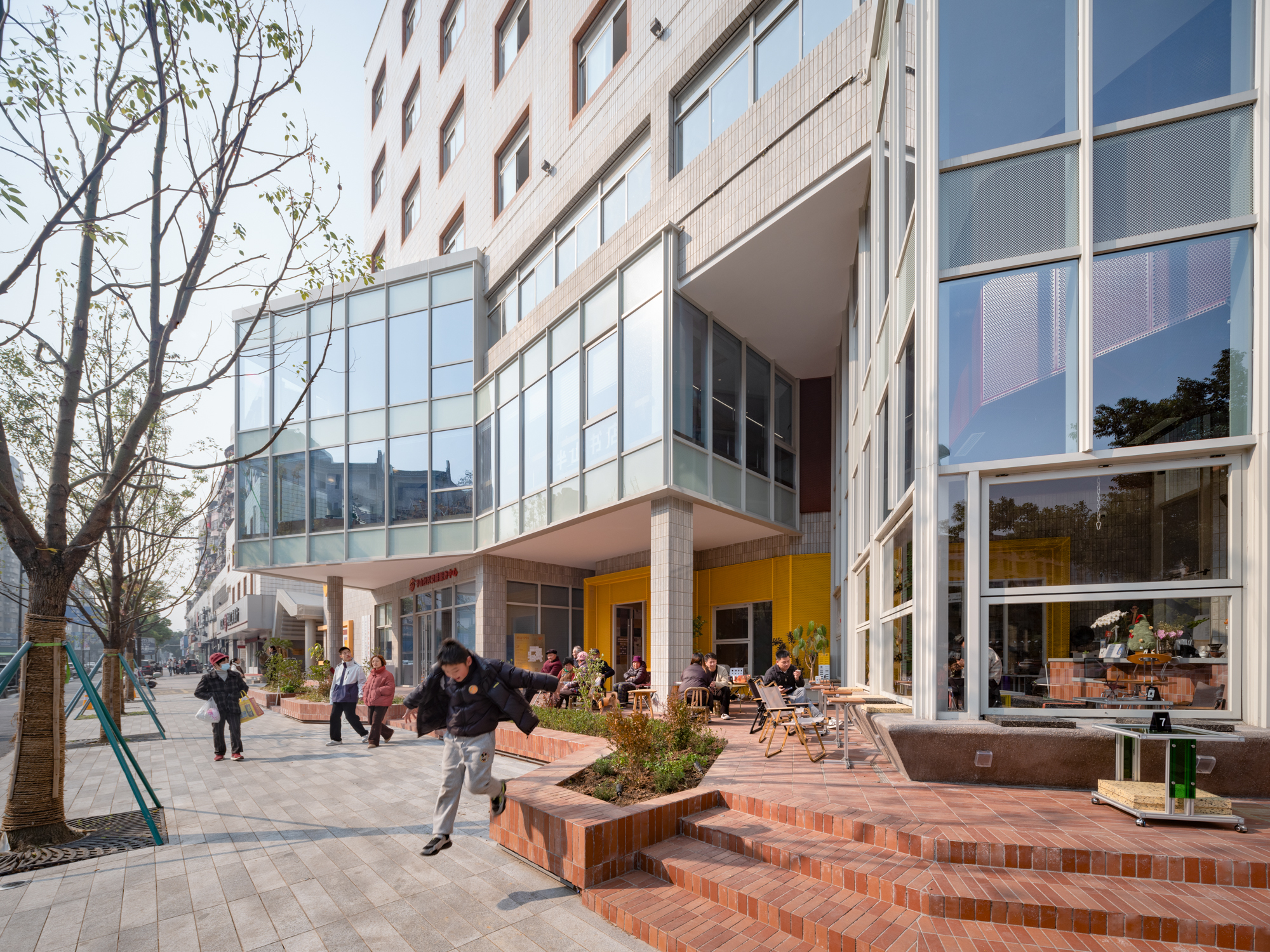 Low angle of the glass corner volume and red brick steps, with people seated at outdoor tables beside a bare tree