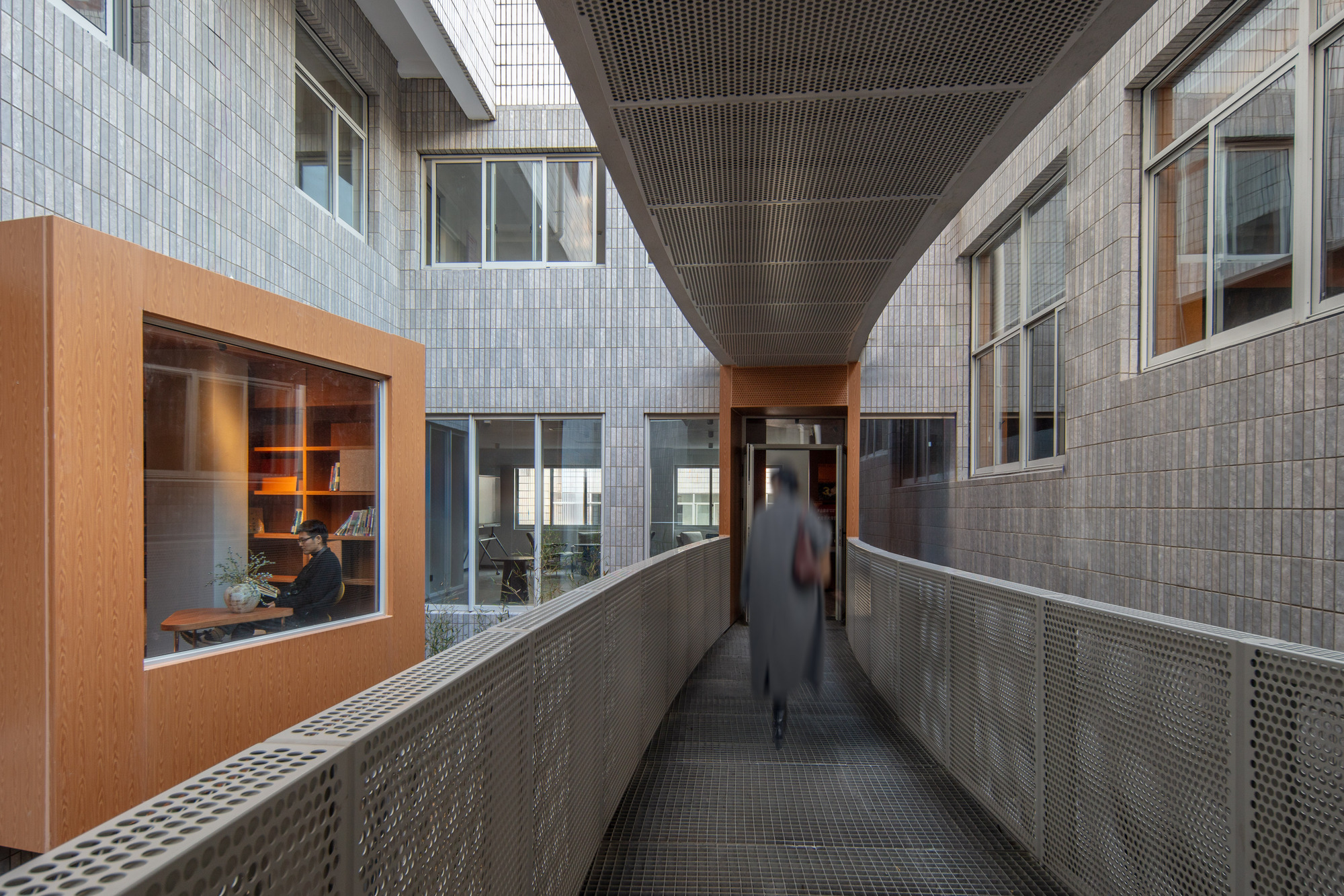 Perforated metal walkway between tiled walls, with a blurred figure walking toward a timber-clad room where someone reads at a desk