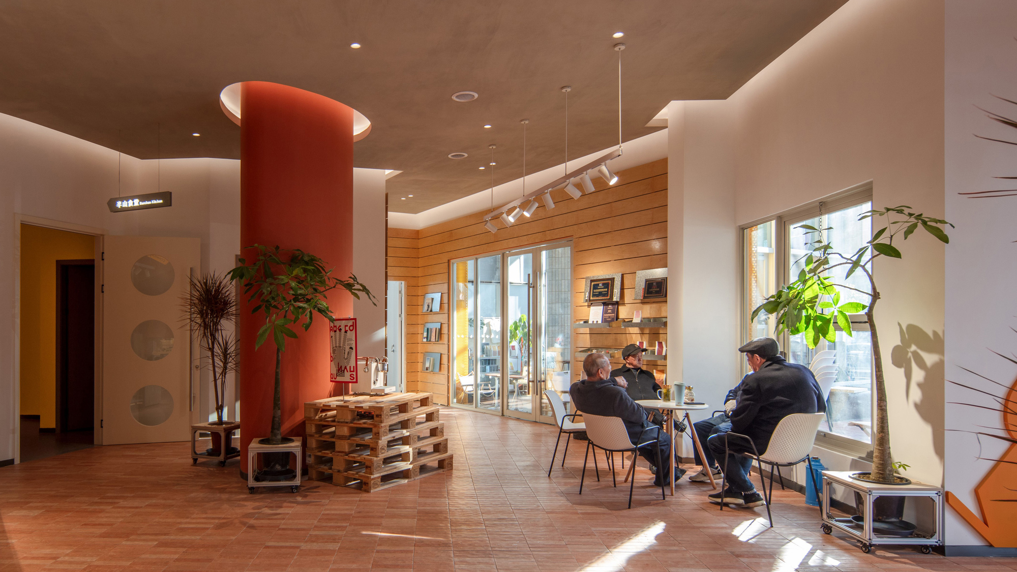 Ground floor lounge with an orange round column and pallet table, where seated men talk in low afternoon sunlight