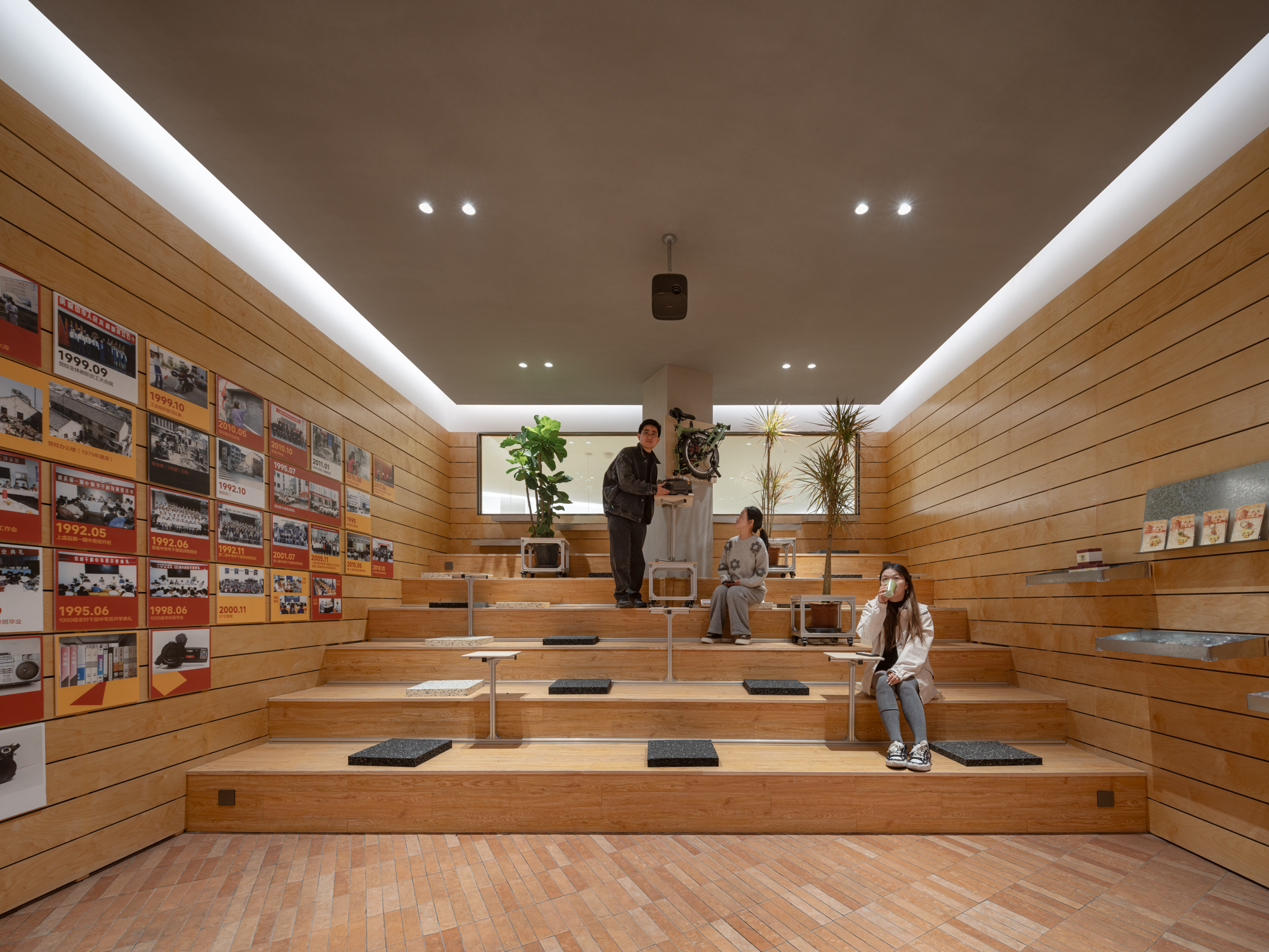 Stepped timber seating tiers lined with framed photographs, with three people and potted plants under a glowing cove-lit ceiling