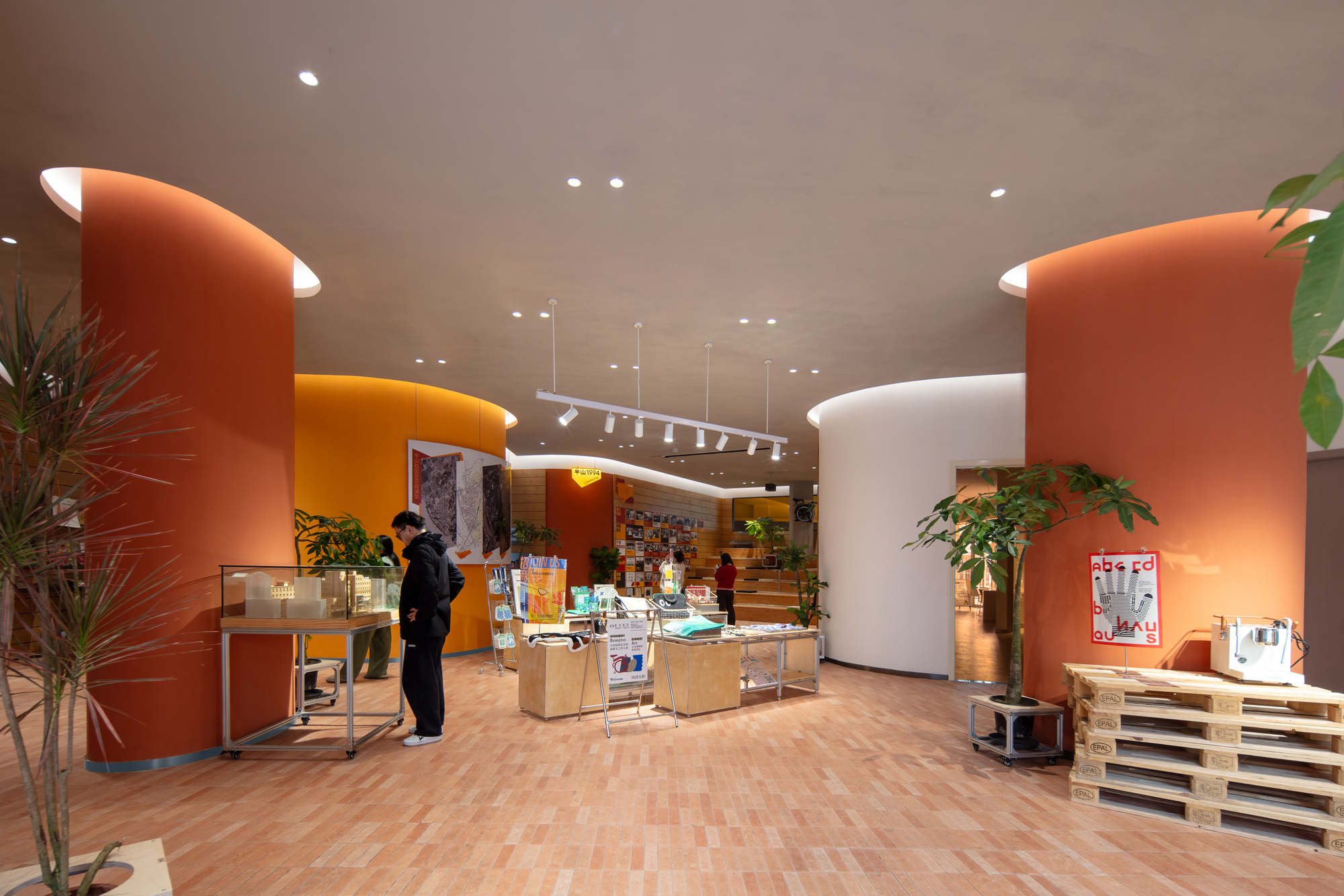 Ground floor retail space with curved orange and white walls, display tables and potted plants, a visitor browsing in warm light
