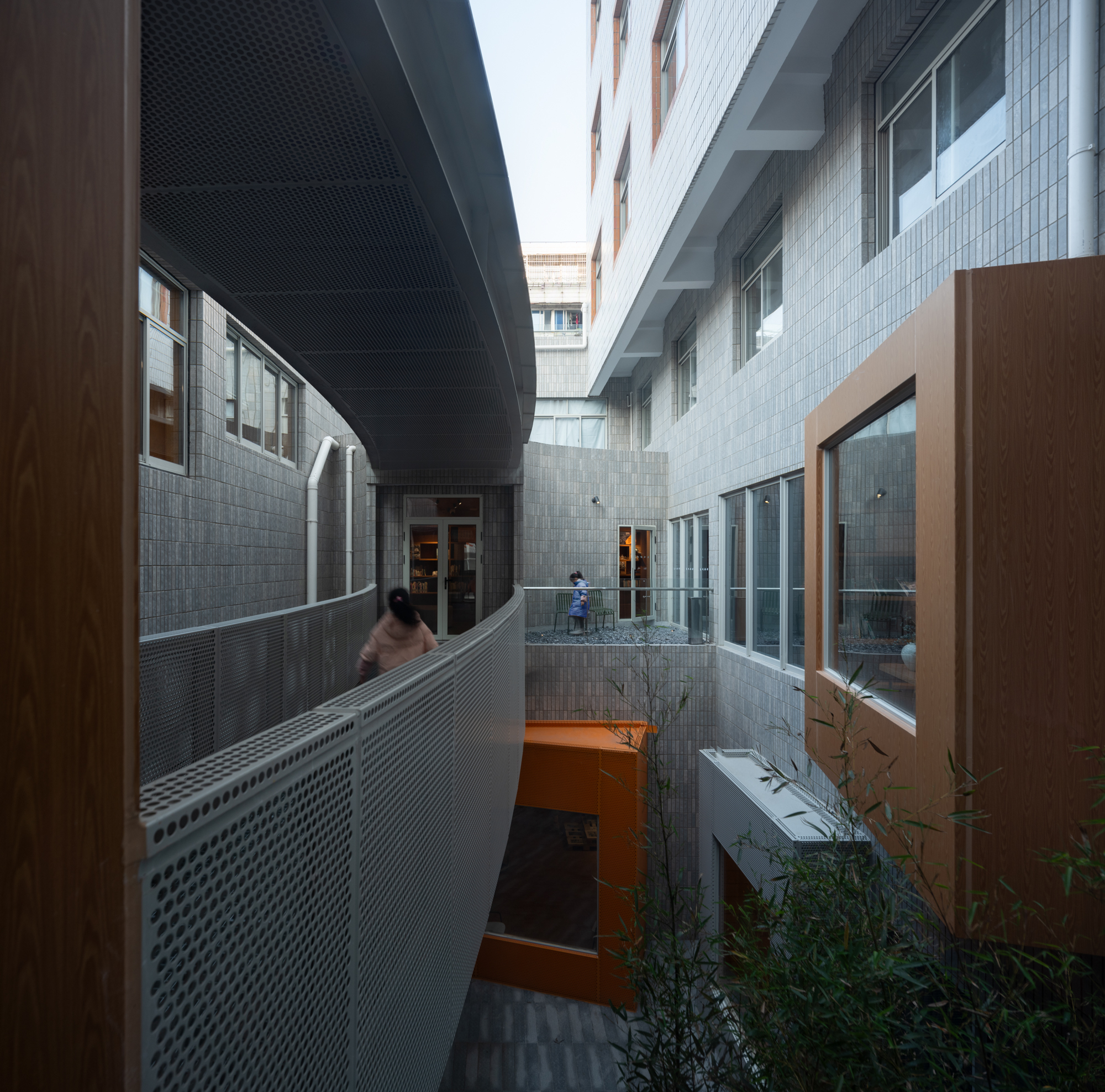 Curving perforated metal walkway between tiled walls and a timber-clad window box, with bamboo in the foreground and a person walking