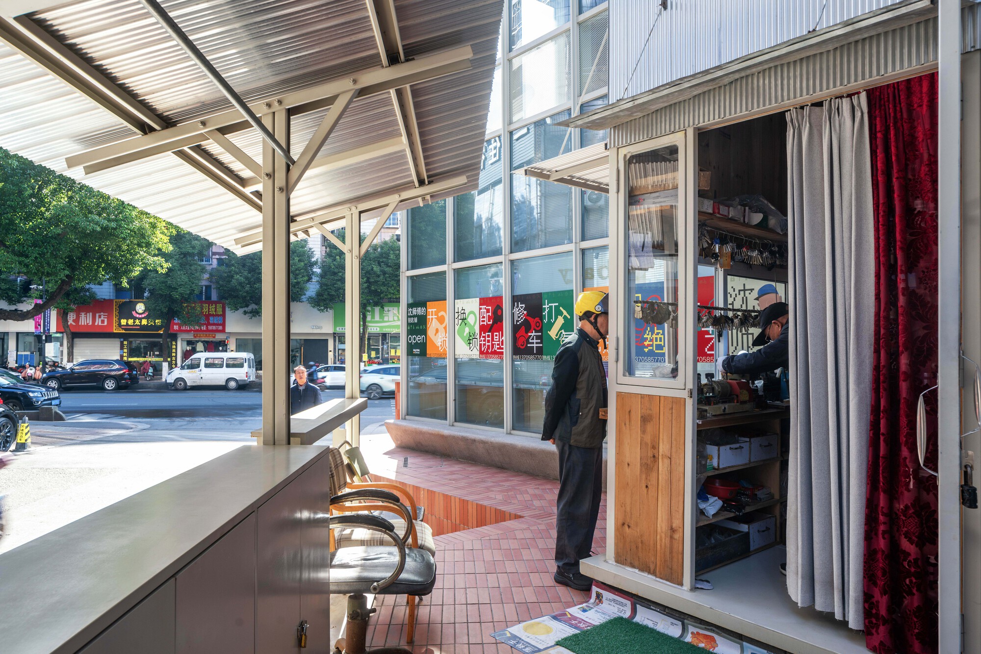 Small corrugated-roof kiosk with a key cutter standing at the counter, red velvet curtain and street traffic beyond in morning sun