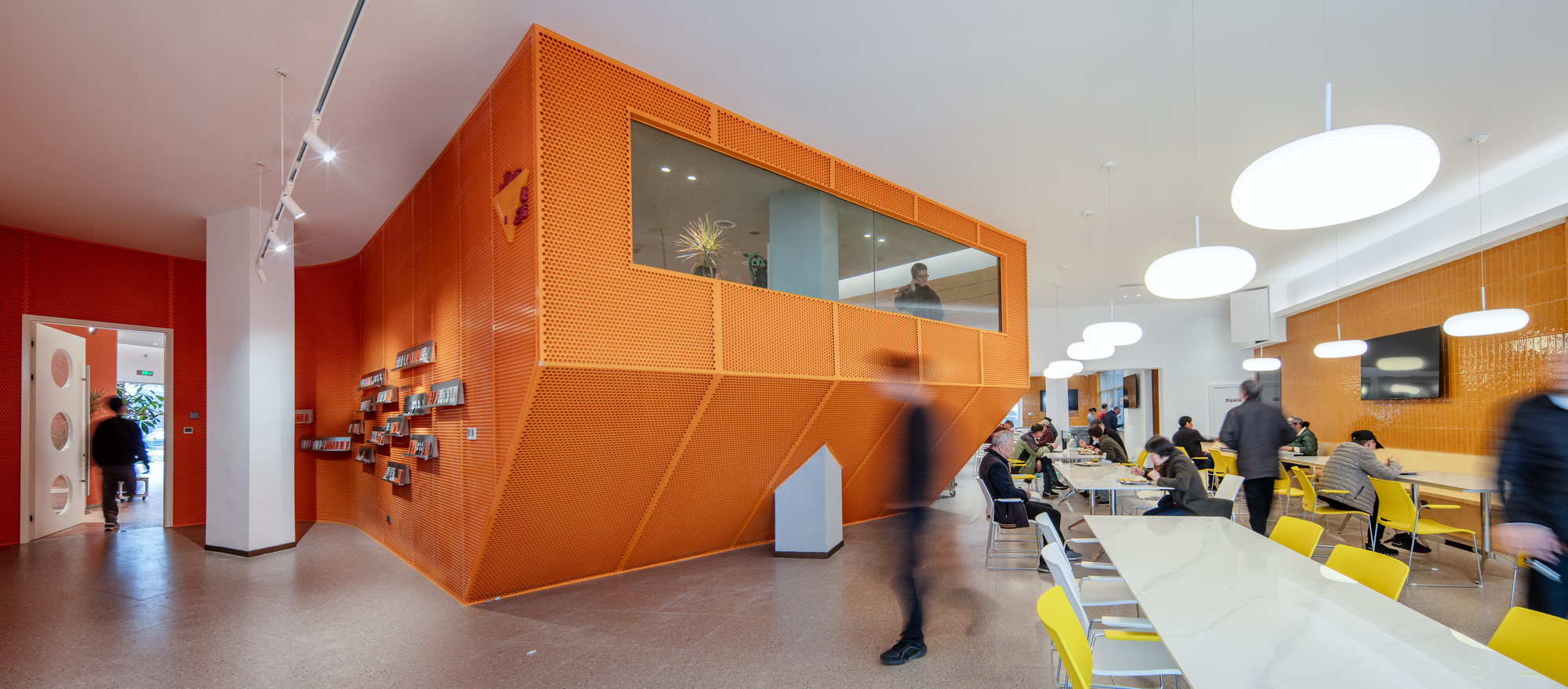 Orange perforated metal volume with angled panels and a window box, set in a reading hall with people passing in motion blur