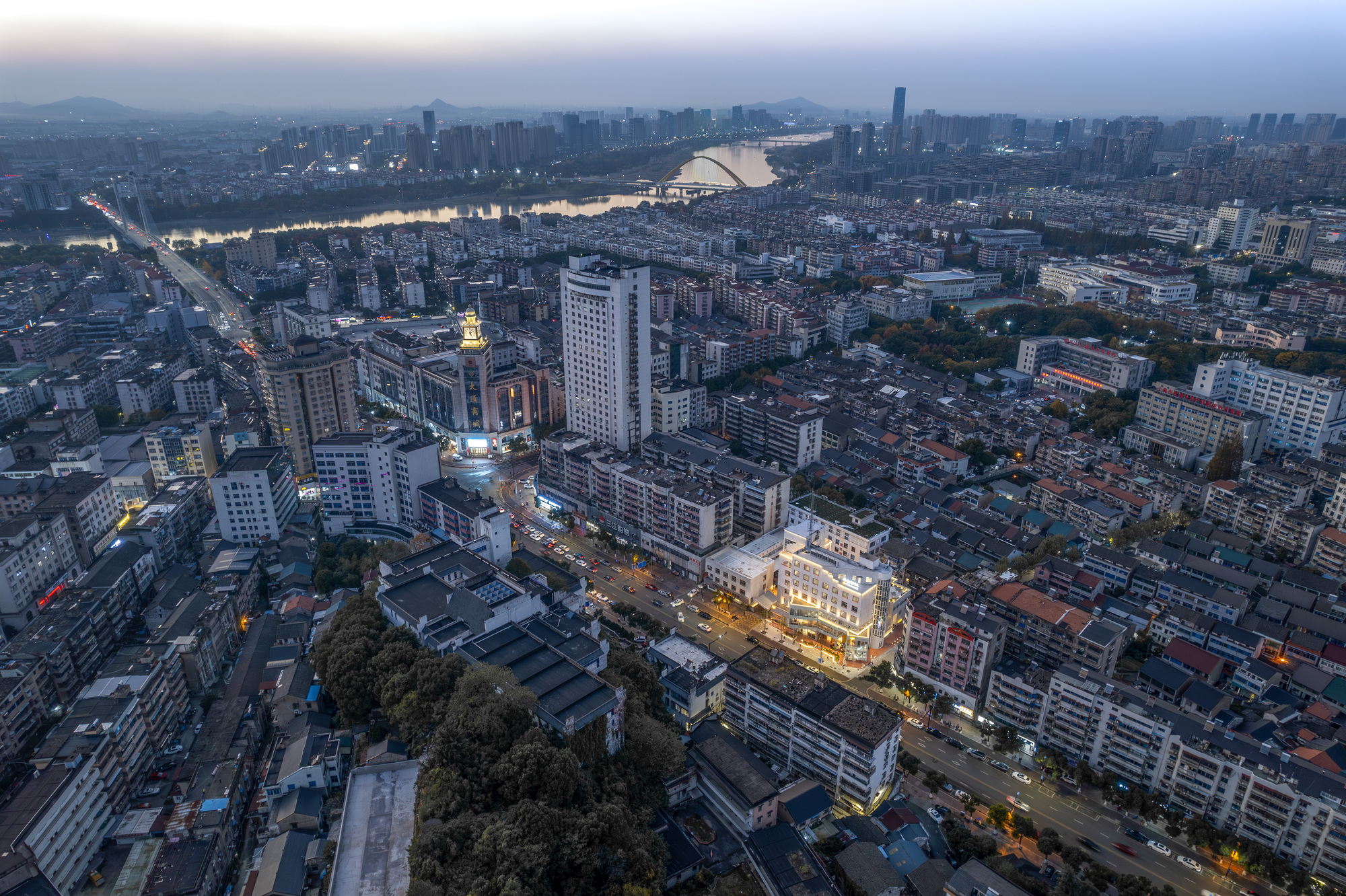 Aerial view of a dense city district at dusk with a river, bridge and a lit low building at the center