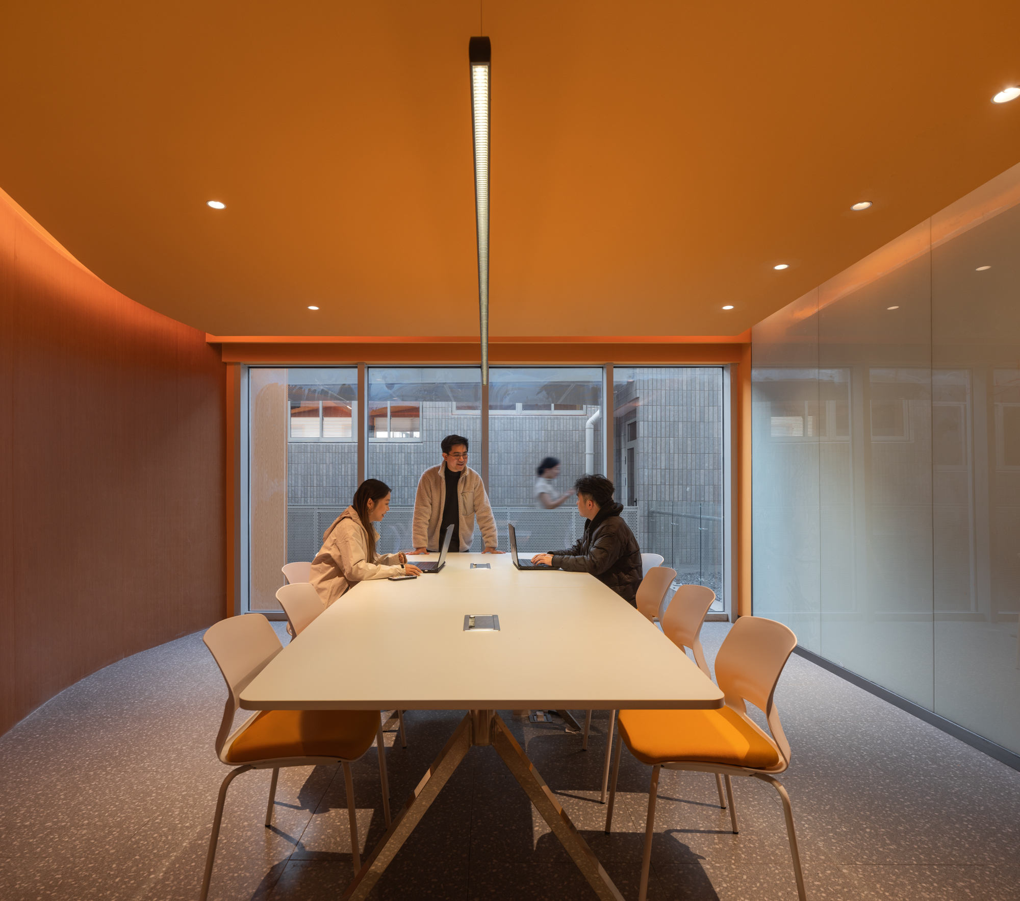Meeting room with a curved orange wall, long white table and three people working at laptops before a tall window