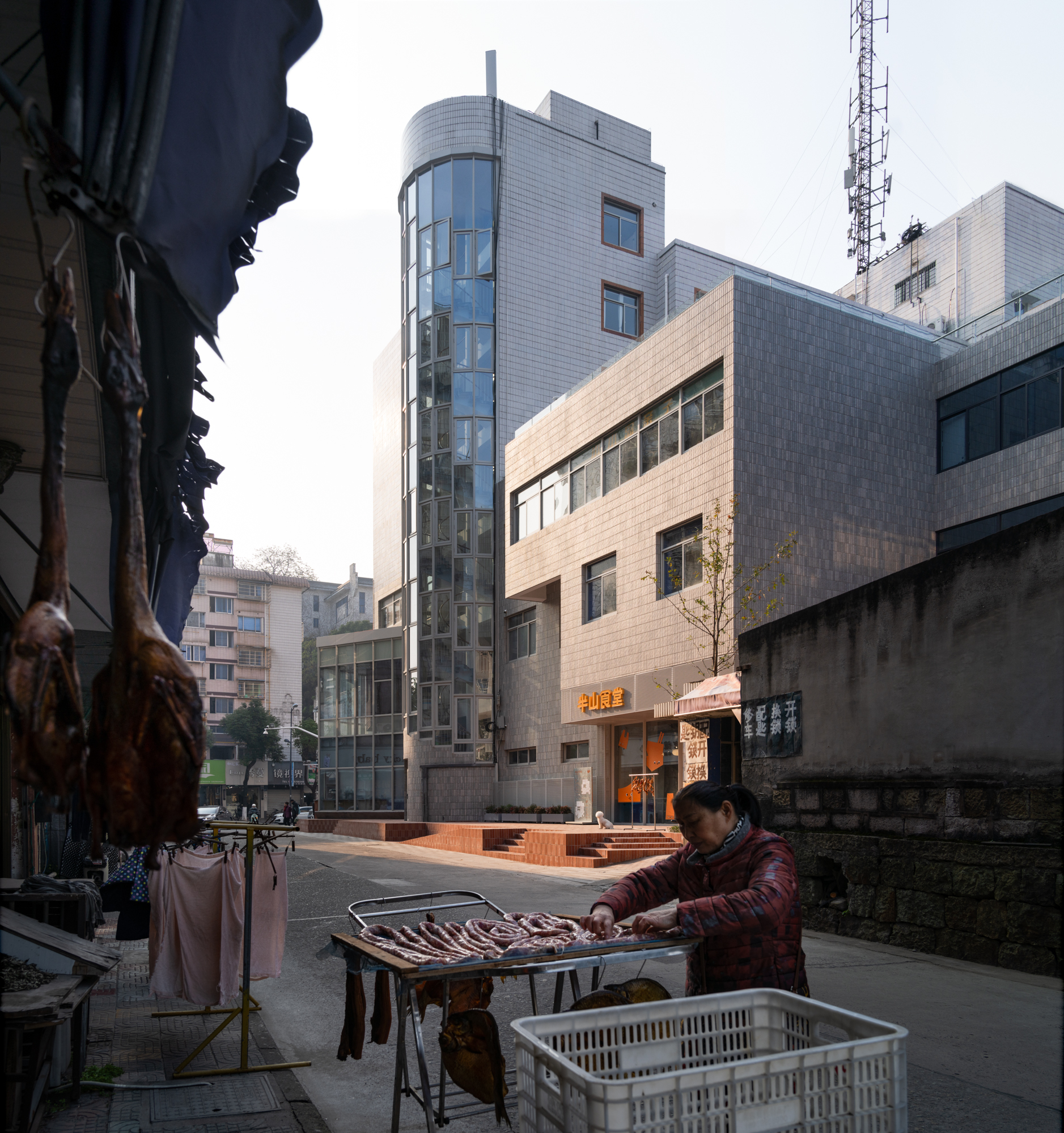 Street view of a gray tiled building with a curved glass tower, a vendor laying out cured meat in the foreground