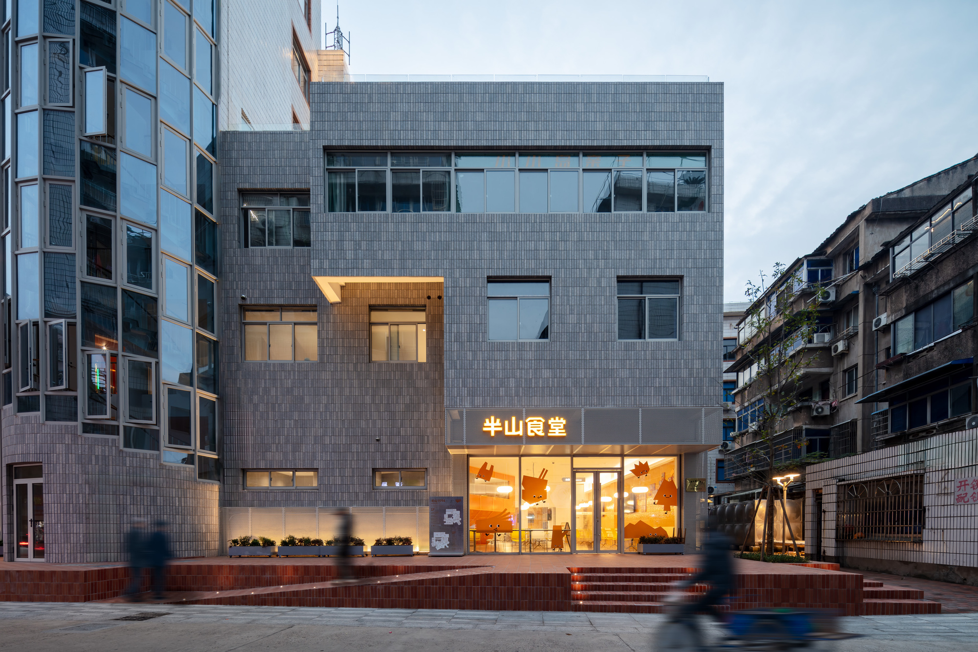 Gray tiled facade with a glowing orange storefront and red stepped terrace at dusk, a cyclist blurring past in front