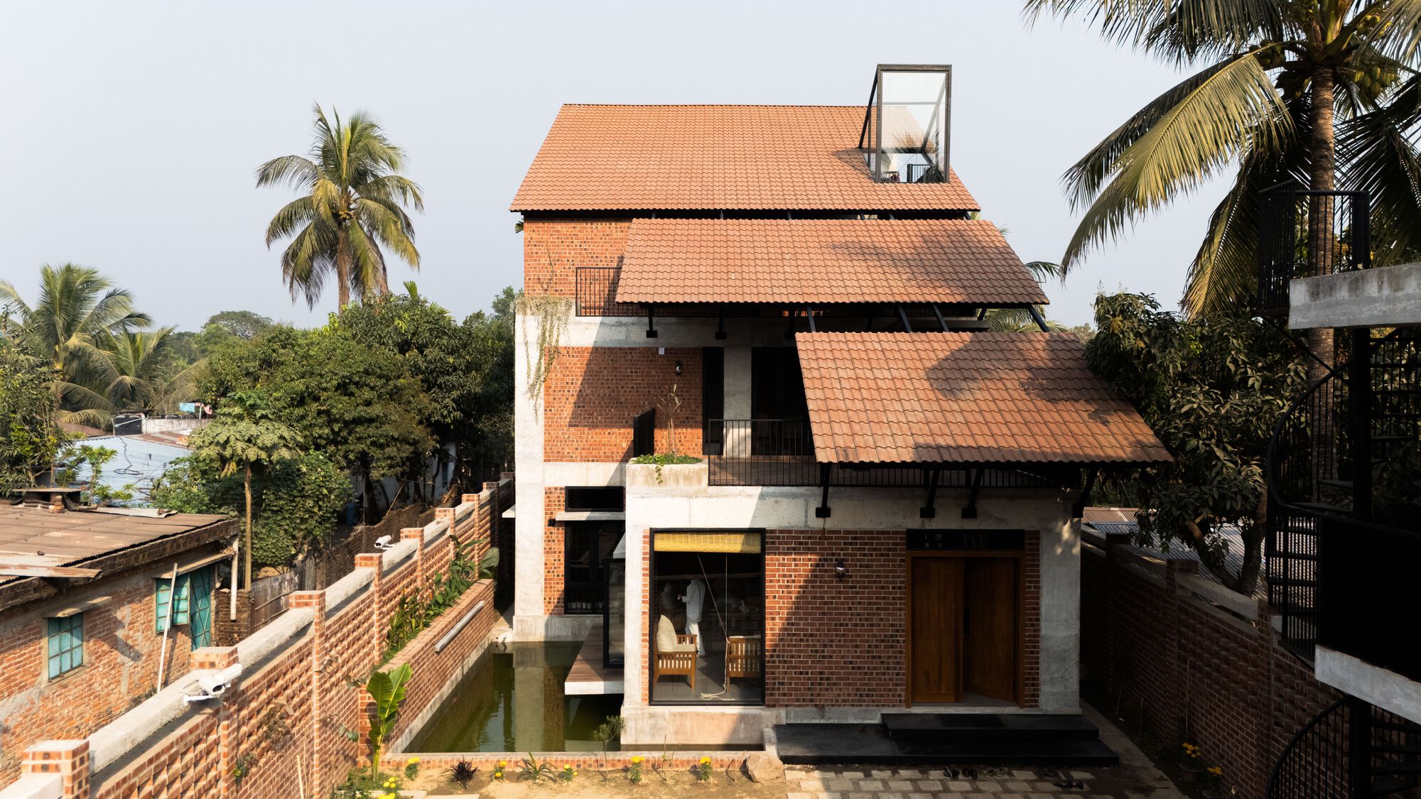 Front facade of stepped terracotta tile roofs over exposed brick and concrete, with a small pond and palm trees in hazy daylight