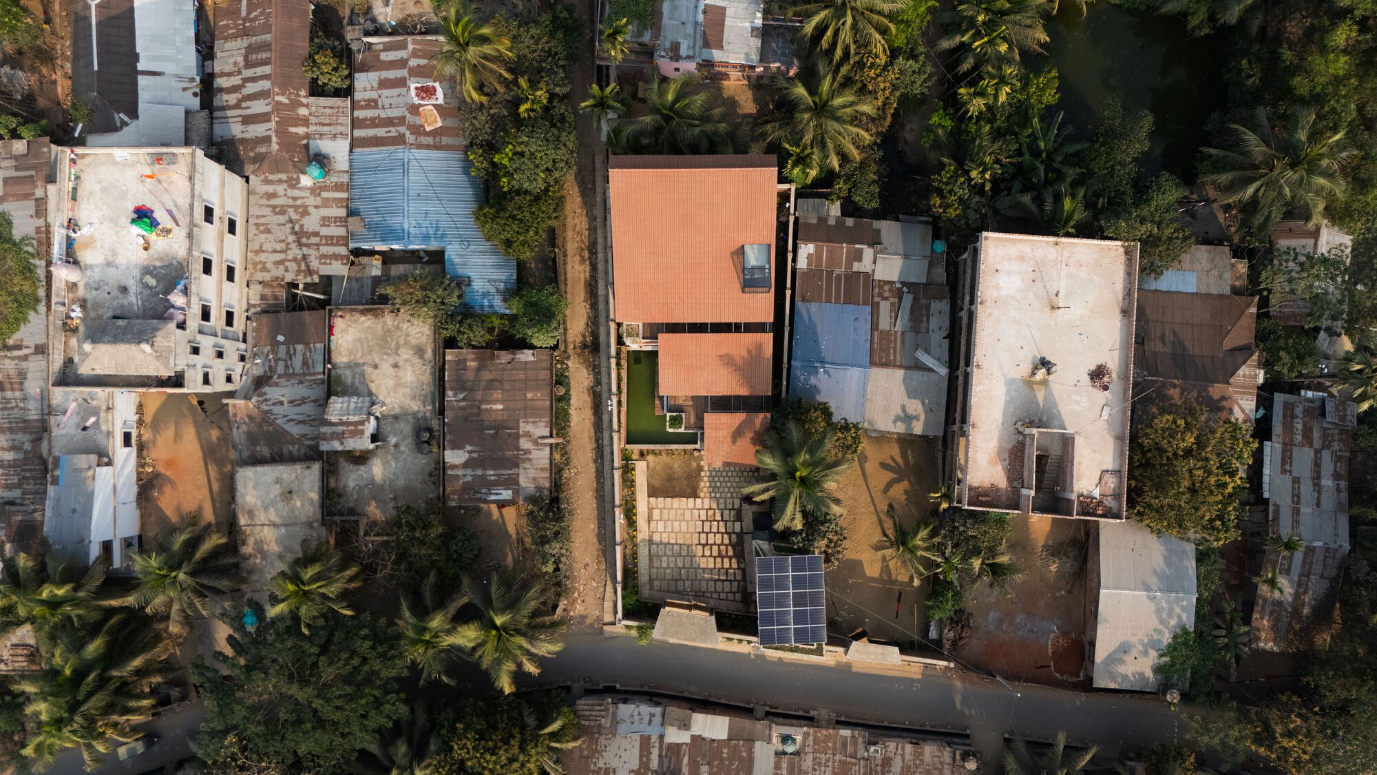 Top-down drone view of the tiled roofs among neighbouring corrugated metal and flat roofs, with palms and a solar panel nearby
