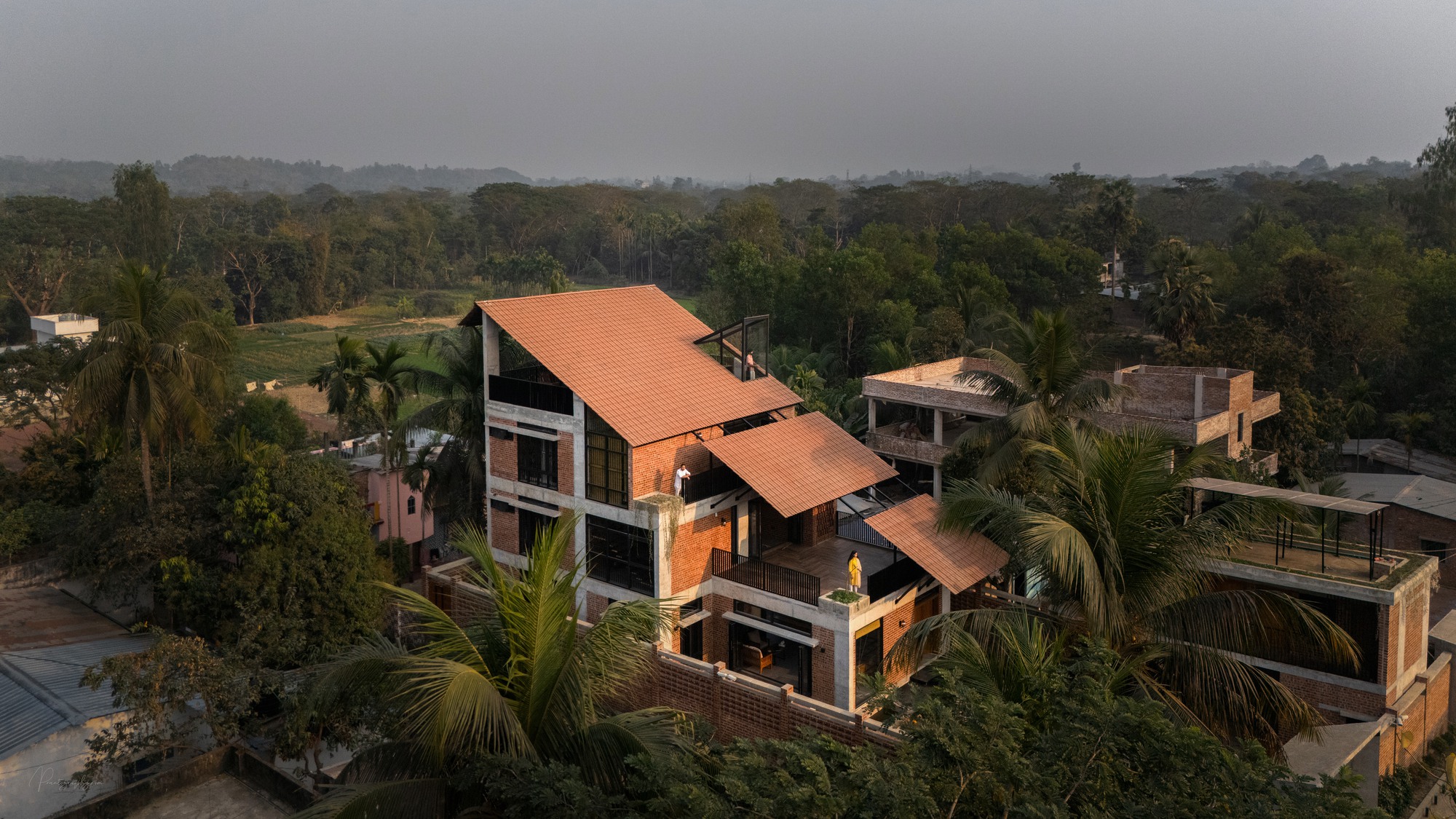 Aerial view of the multi-level brick house with layered tile roofs and a person on the terrace, surrounded by palms at dusk