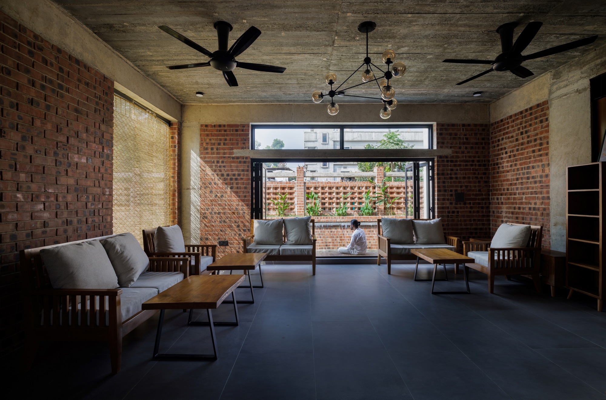 Living room with exposed concrete ceiling, brick walls and timber sofas, a seated person facing the open window and perforated brick screen