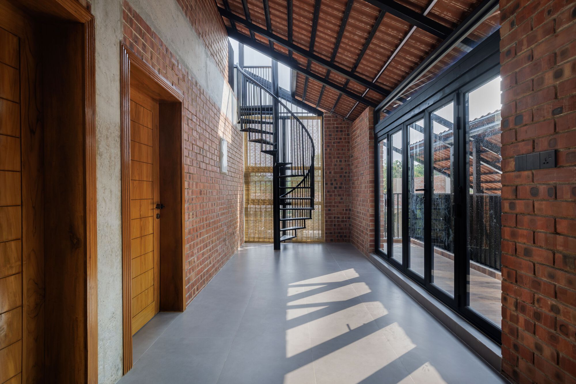 Corridor with timber doors, brick walls and a black spiral staircase, and sunlight from the glazed wall striping the grey floor
