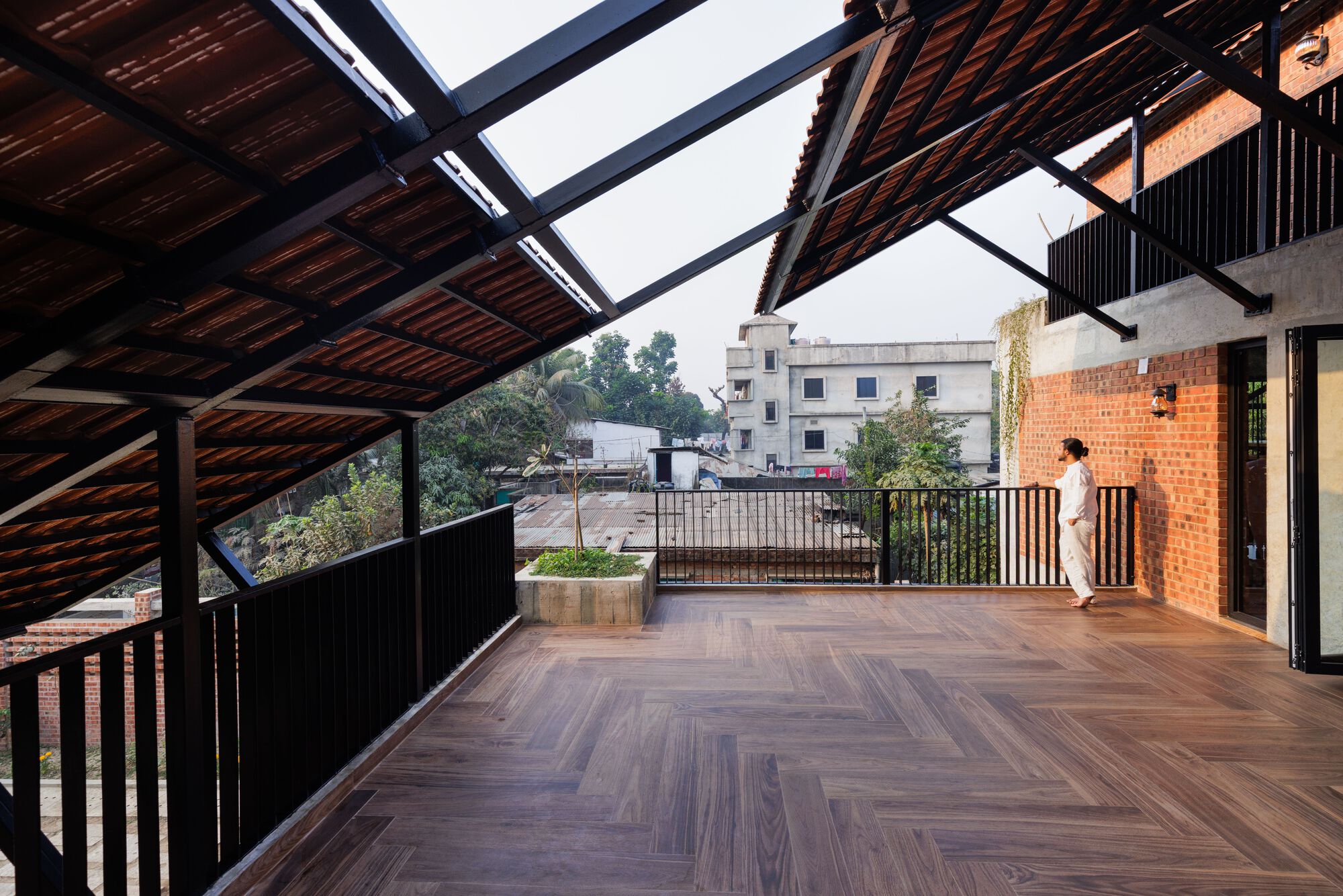 Covered terrace with herringbone timber floor, steel-framed tile roof and black railings, a person looking out over neighbouring buildings in soft light