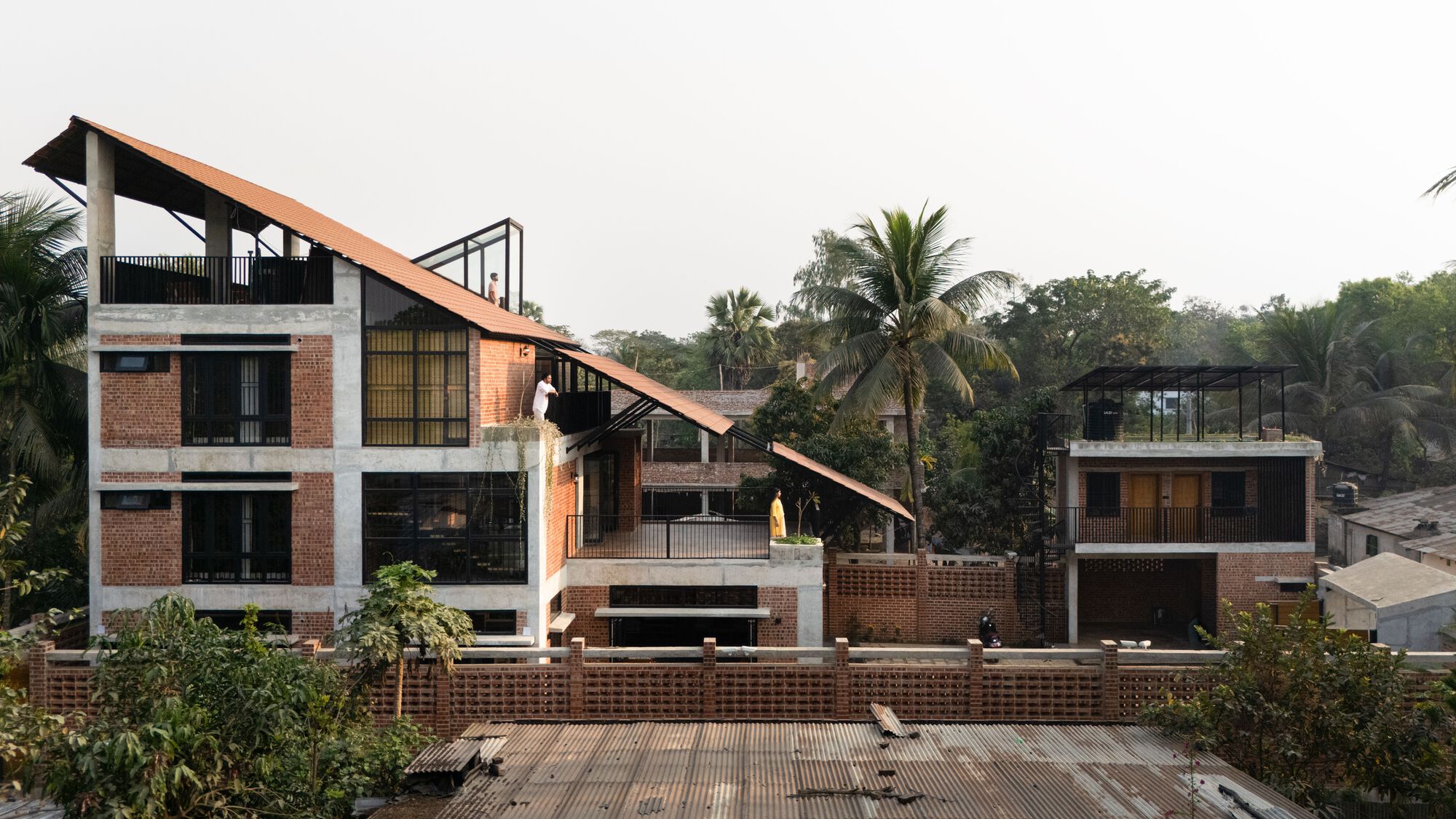Street-side view of the brick and concrete house with its sloping terracotta roof, framed by palms in hazy light