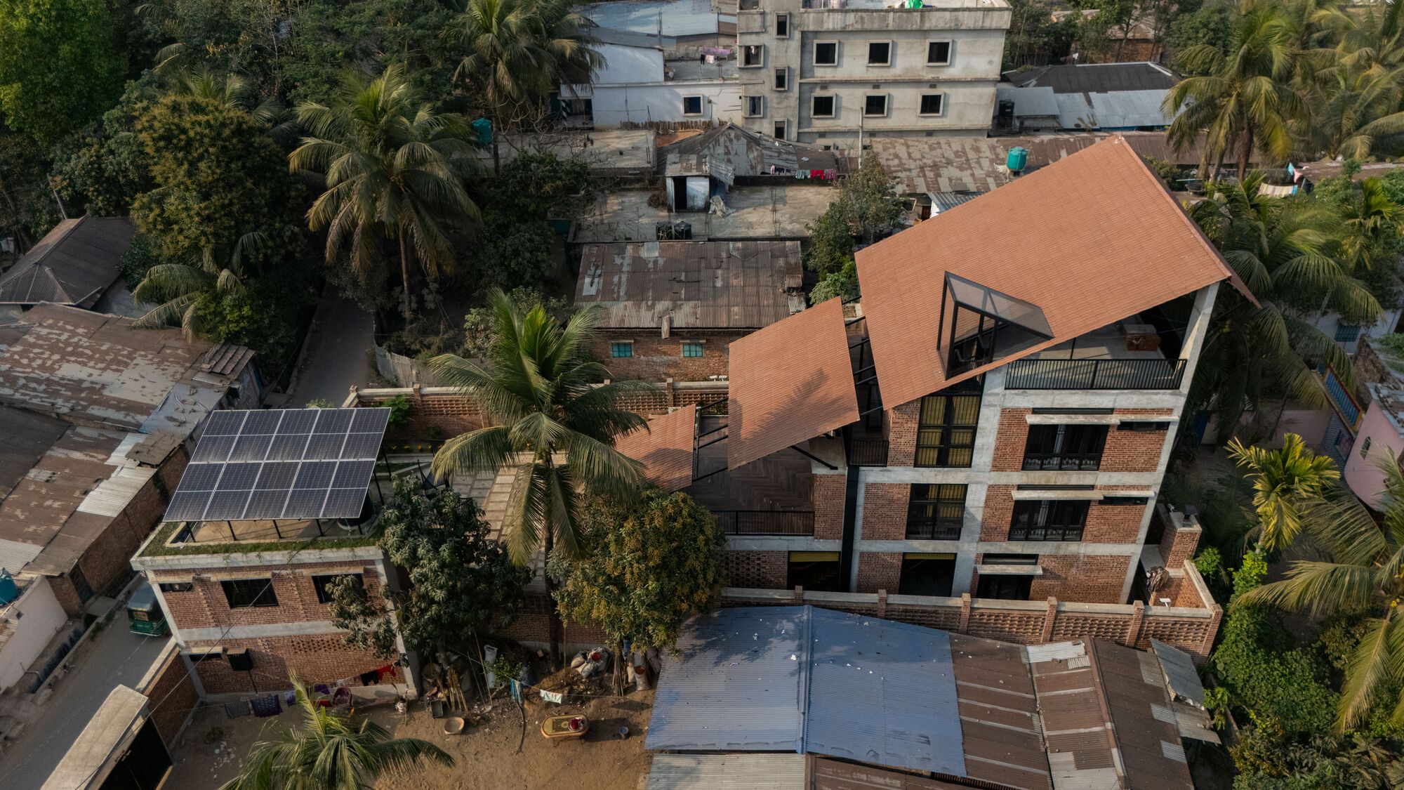 Drone view of the tiled roof planes and brick screen wall among palm trees and neighboring tin roofs