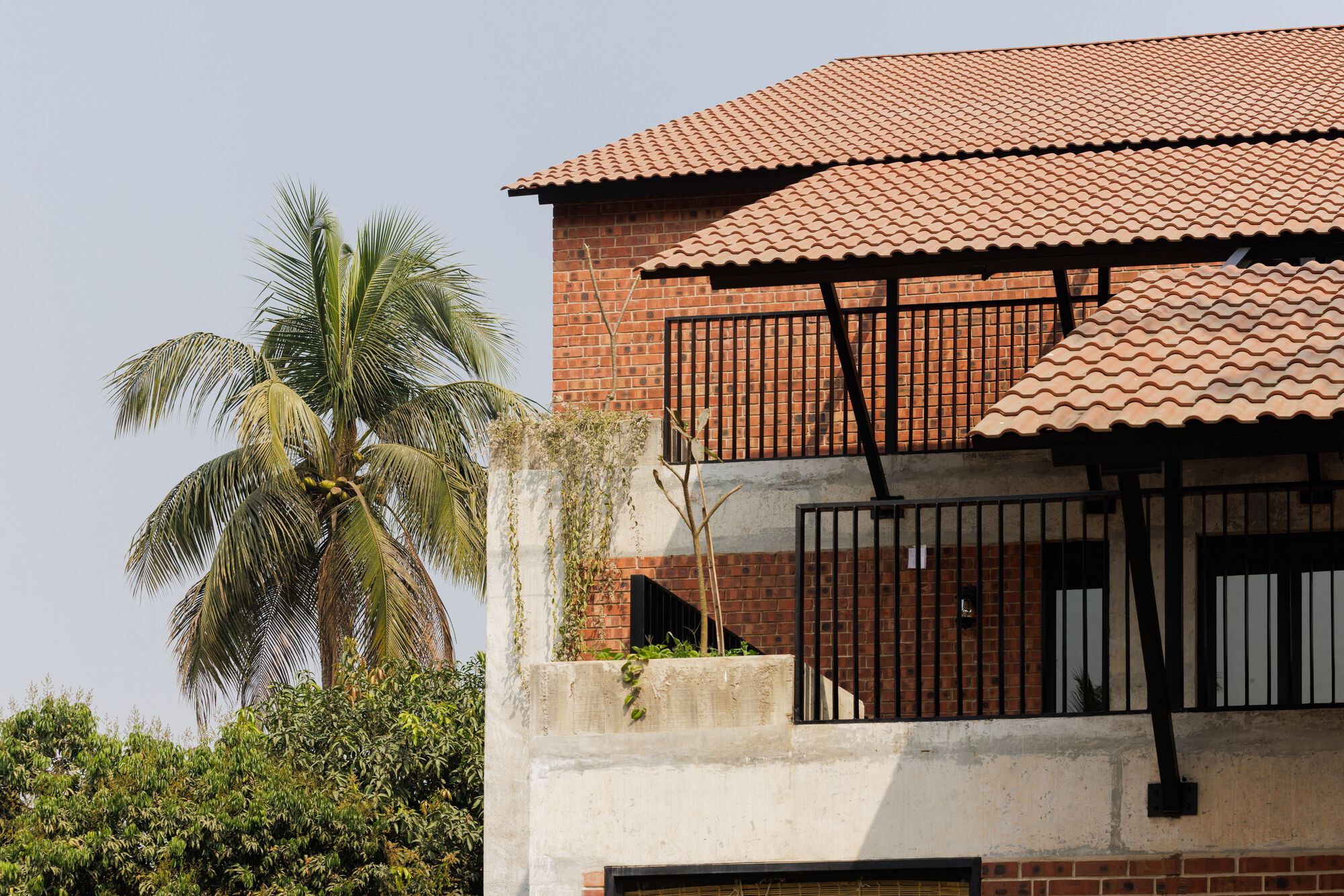 Corner of the brick and concrete volume with overlapping tiled canopies, black railings and a planter, beside a tall palm