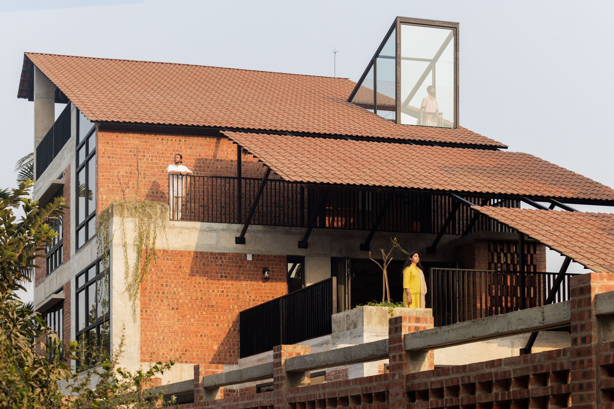 Tiered terracotta roofs on slim steel struts above a brick screen wall, with two people standing on the terraces