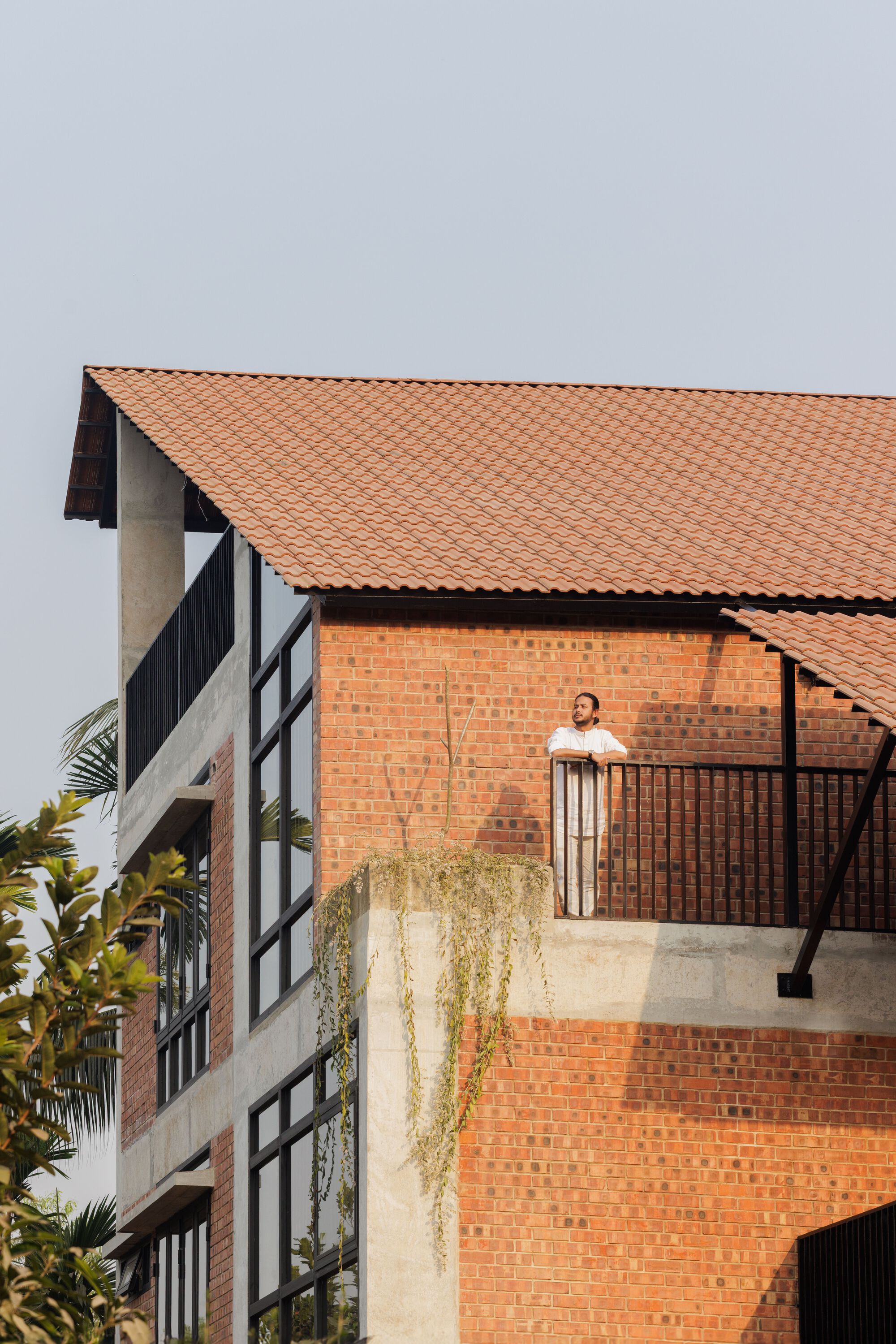 Man leaning on a black balcony railing against a brick wall, with trailing vines and a pitched tile roof above
