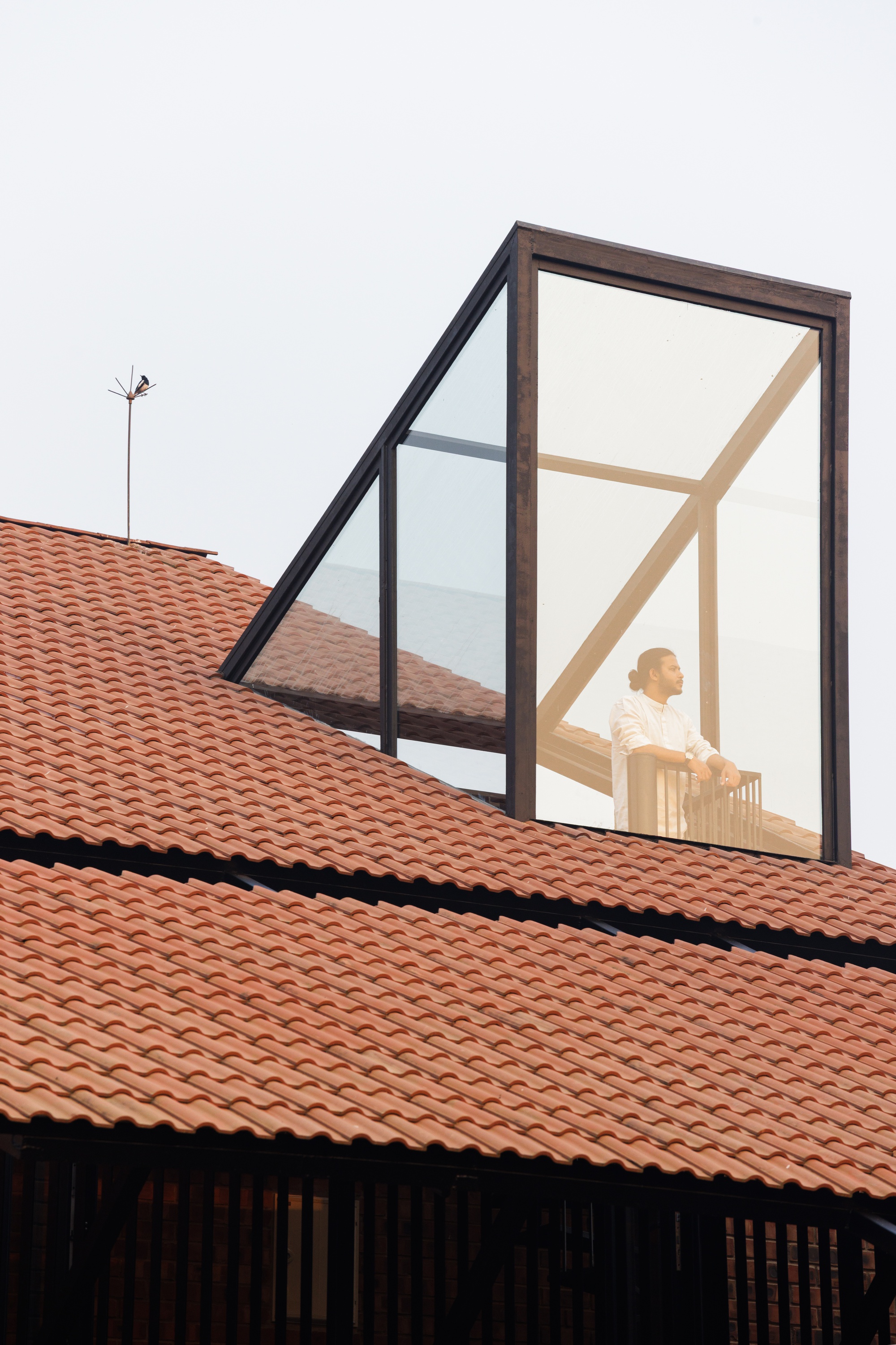 Glazed rooftop dormer with a timber frame rising above terracotta tiles, a person visible inside under a pale hazy sky