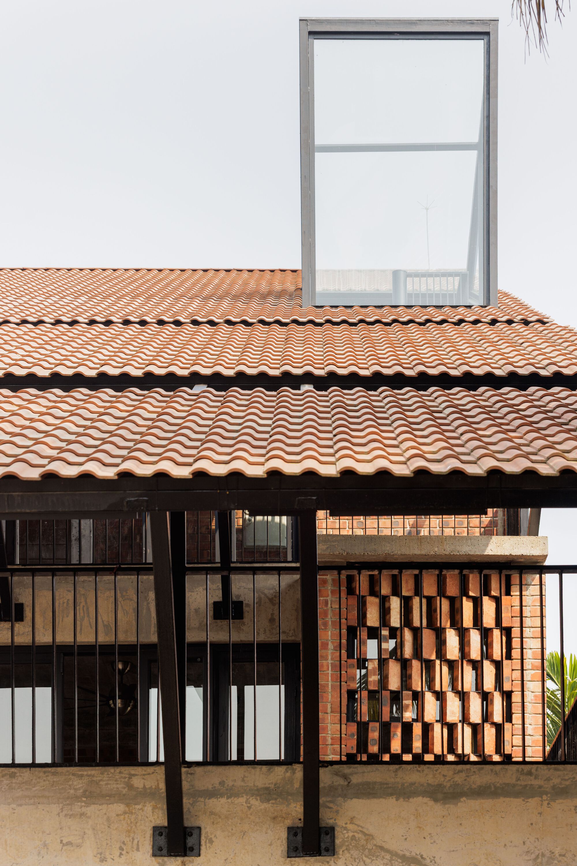 Terracotta tiled roof layered above a steel-framed balcony and a checkerboard brick screen, with a glazed box rising against a pale sky