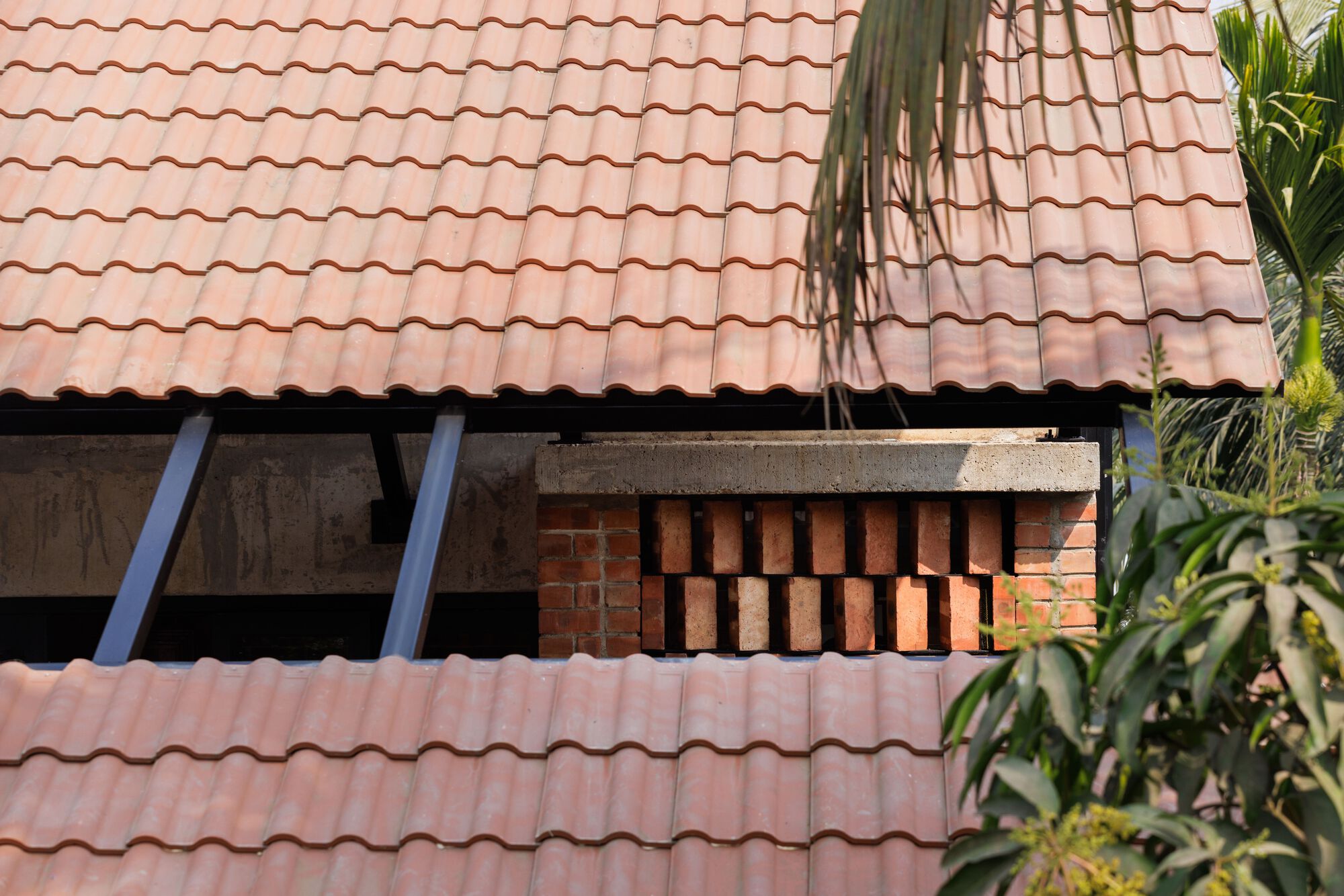 Close view of a perforated brick panel under the clay tile roof, framed by palm fronds and leafy shrubs