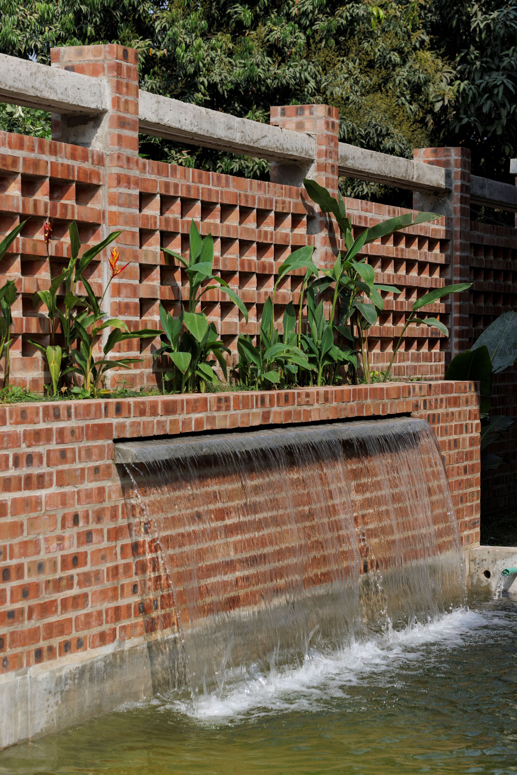 Water spilling over a brick weir below a perforated brick wall planted with heliconia and broad-leaved plants