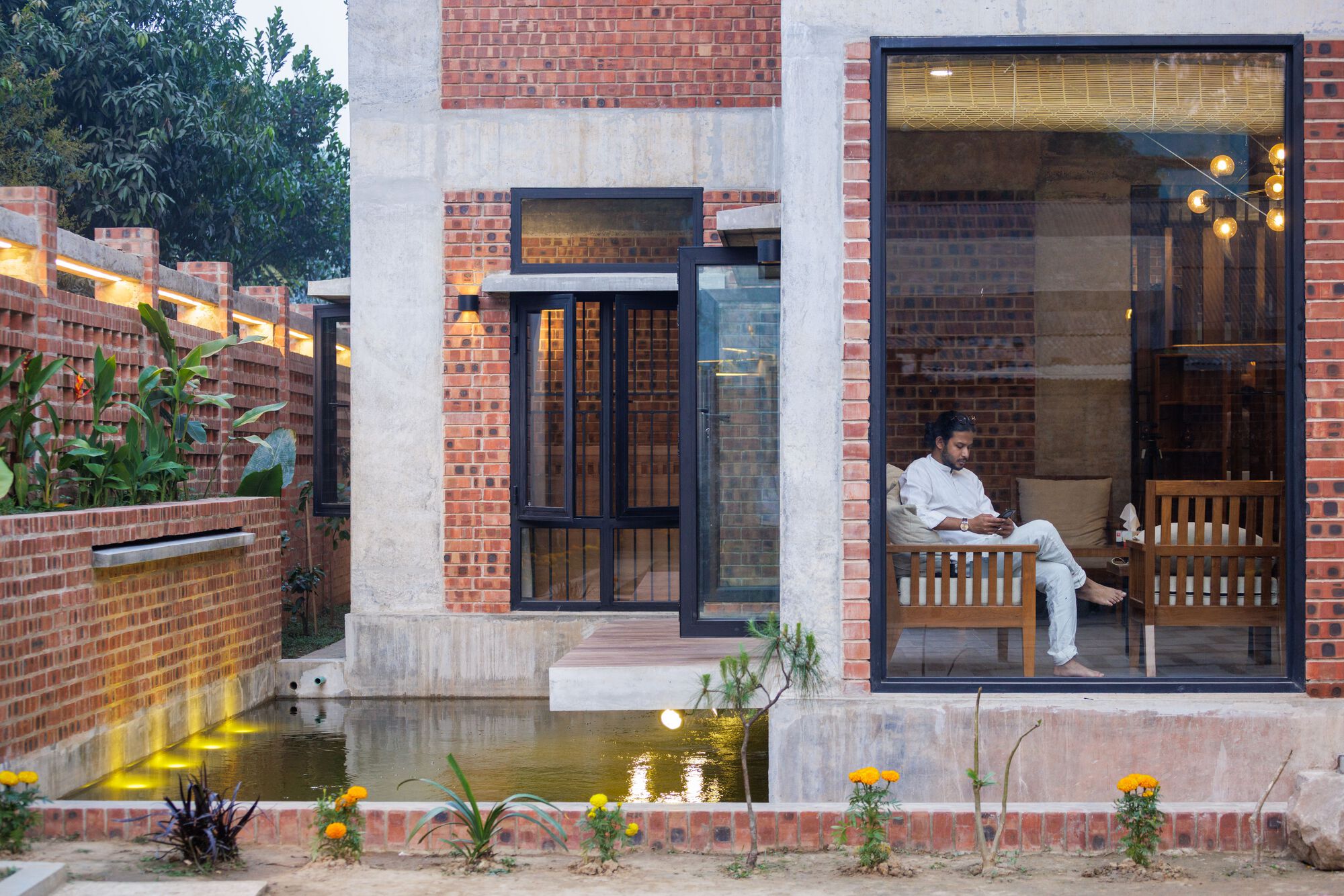 A man seated in an armchair behind a large steel-framed window at dusk, with lit pond edge and marigolds outside