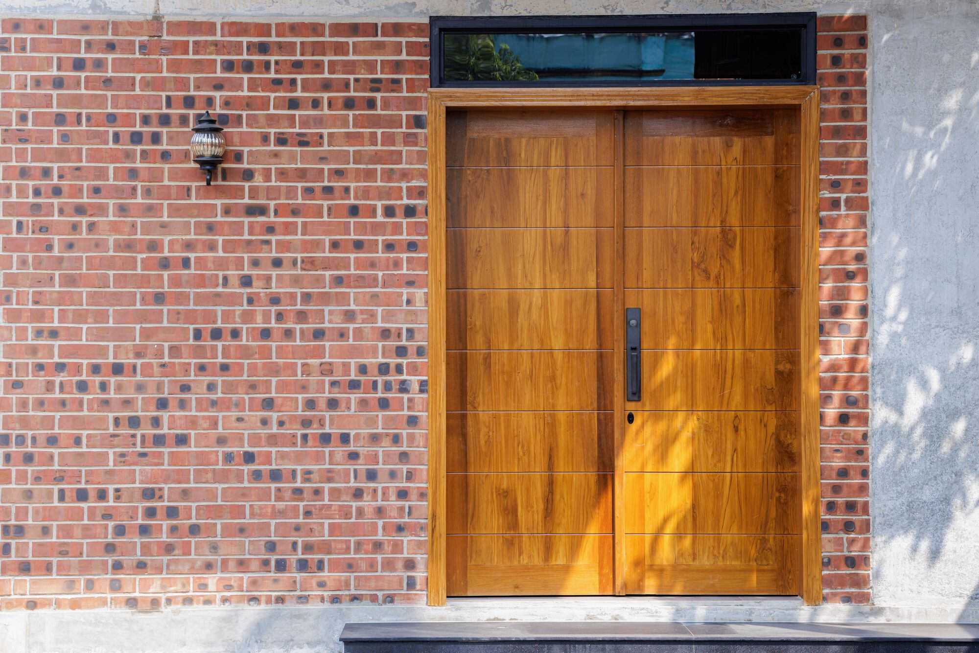 Double timber door with a vertical handle set in a brick and concrete wall, dappled with leaf shadows