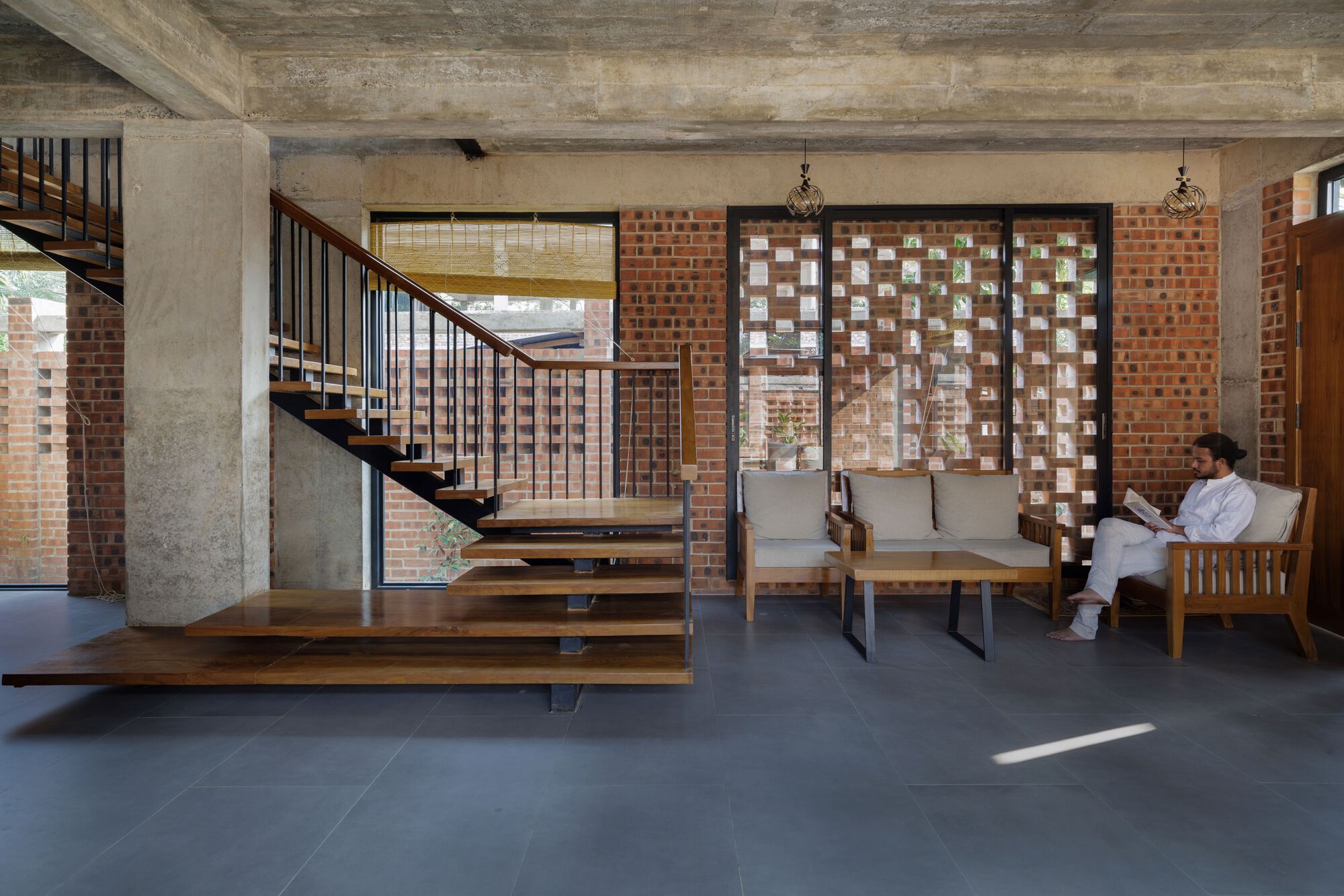 Living area with cantilevered timber stair, perforated brick screen and a man reading in a wooden armchair as a sunbeam crosses the floor
