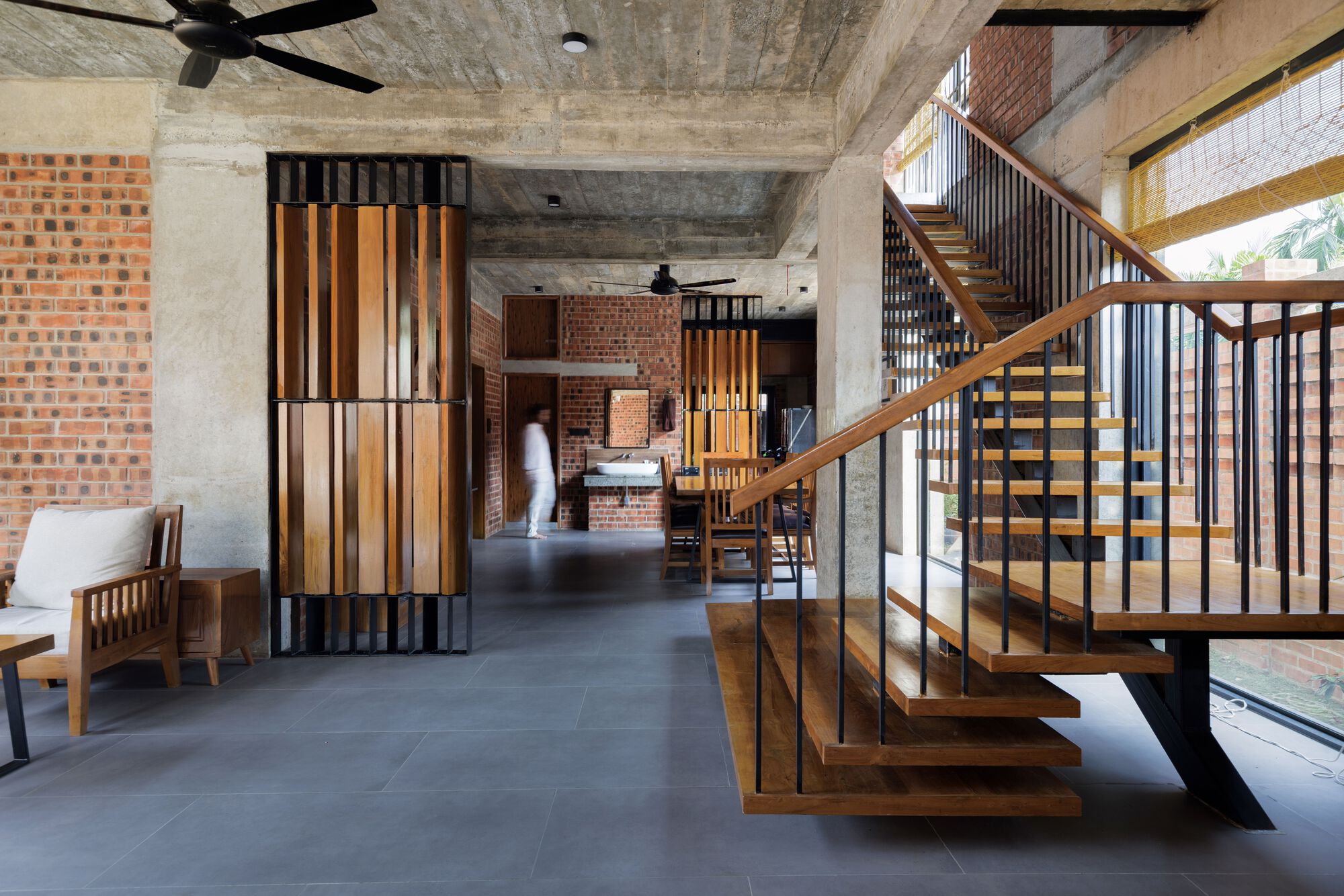 Open ground floor with timber slat partition, floating wooden stair and exposed concrete beams, a person in white walking toward the dining area