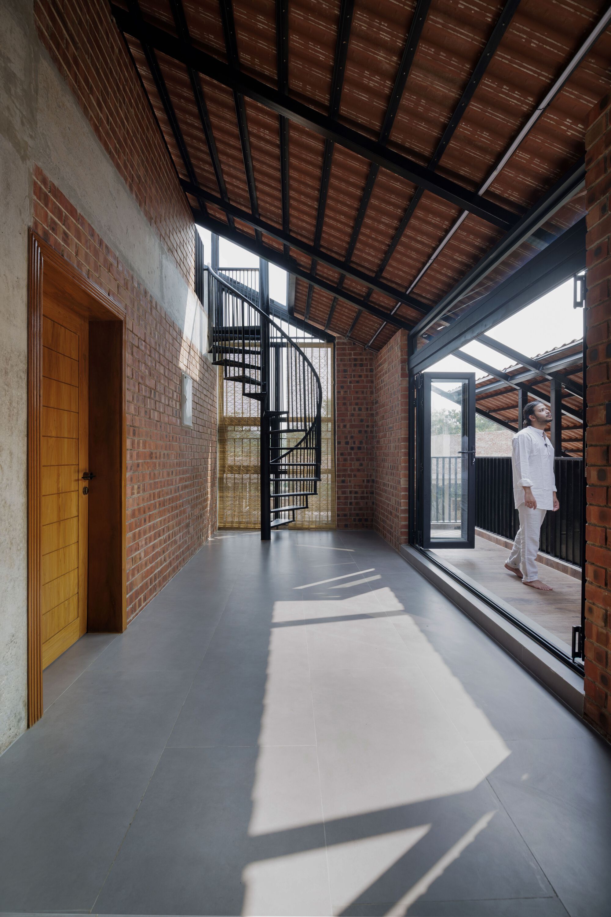 Upper corridor with brick wall, spiral steel stair and tiled pitched roof, afternoon sun casting bright shapes across the grey floor
