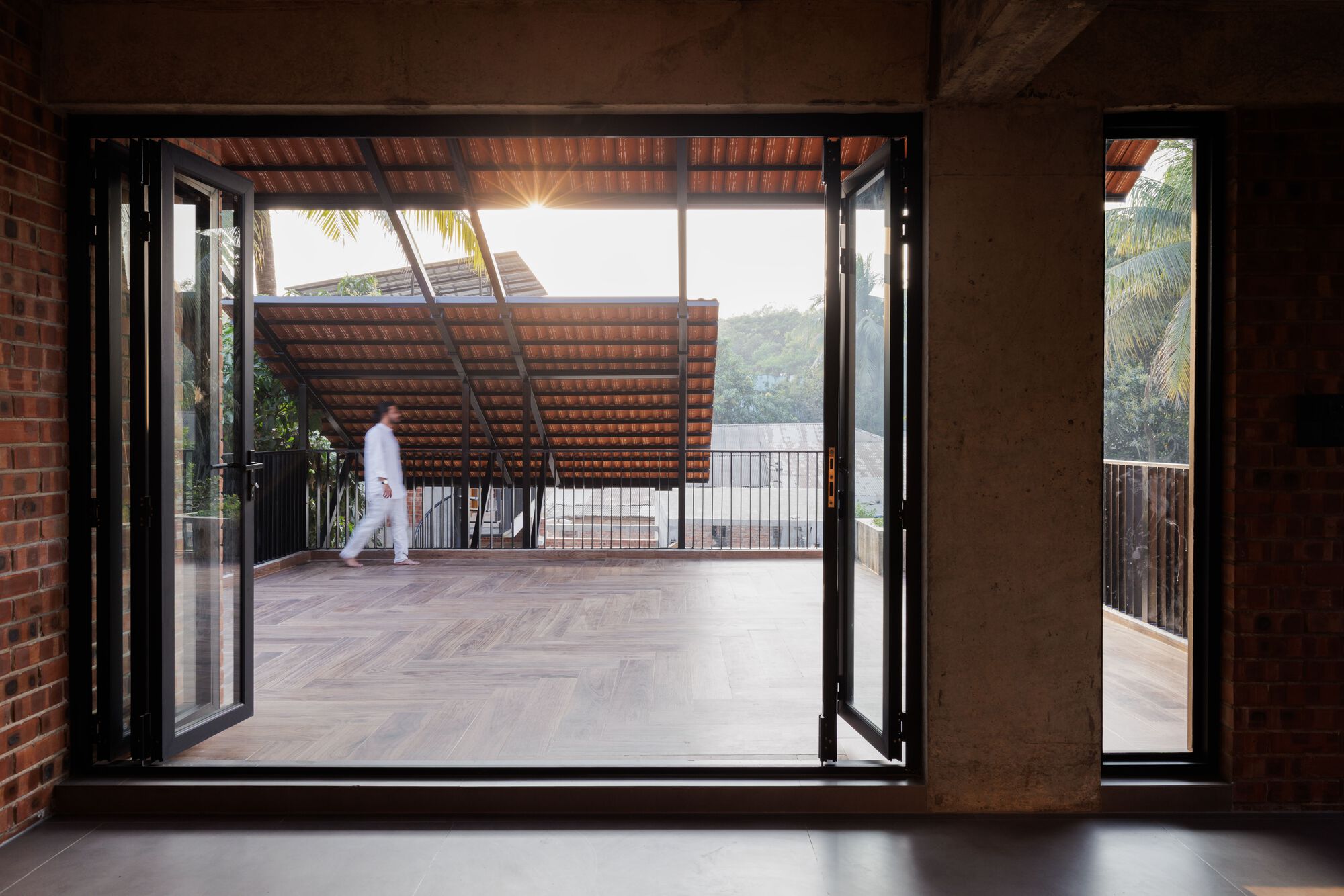 Folding glass doors open to a herringbone timber terrace with a tiled roof, a man walking past at sunset among palms