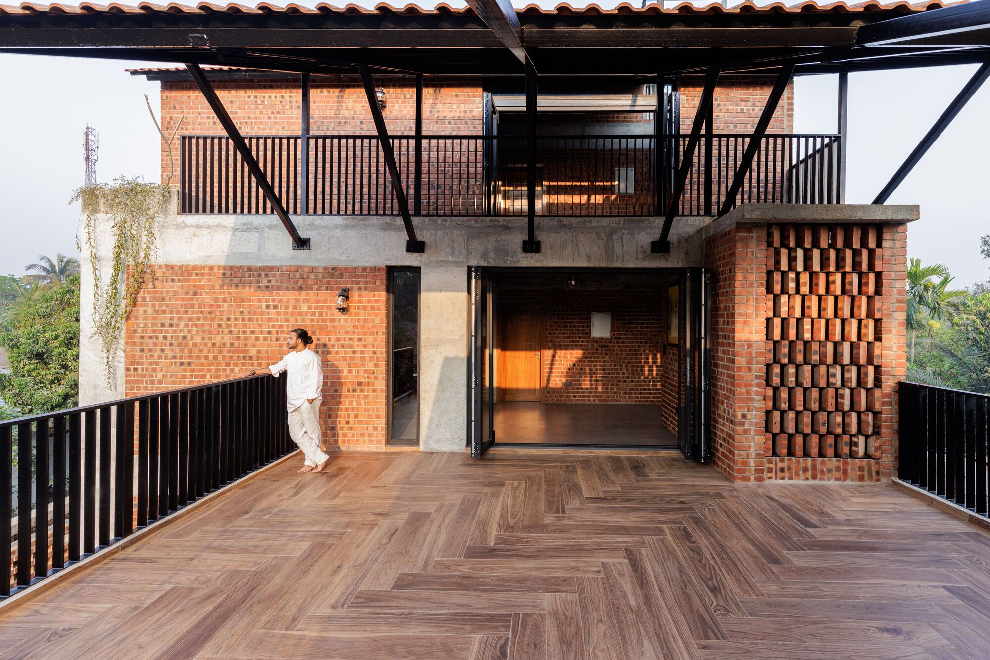 Herringbone timber terrace in front of a brick upper floor with perforated brick panel, steel struts and a man leaning on the railing
