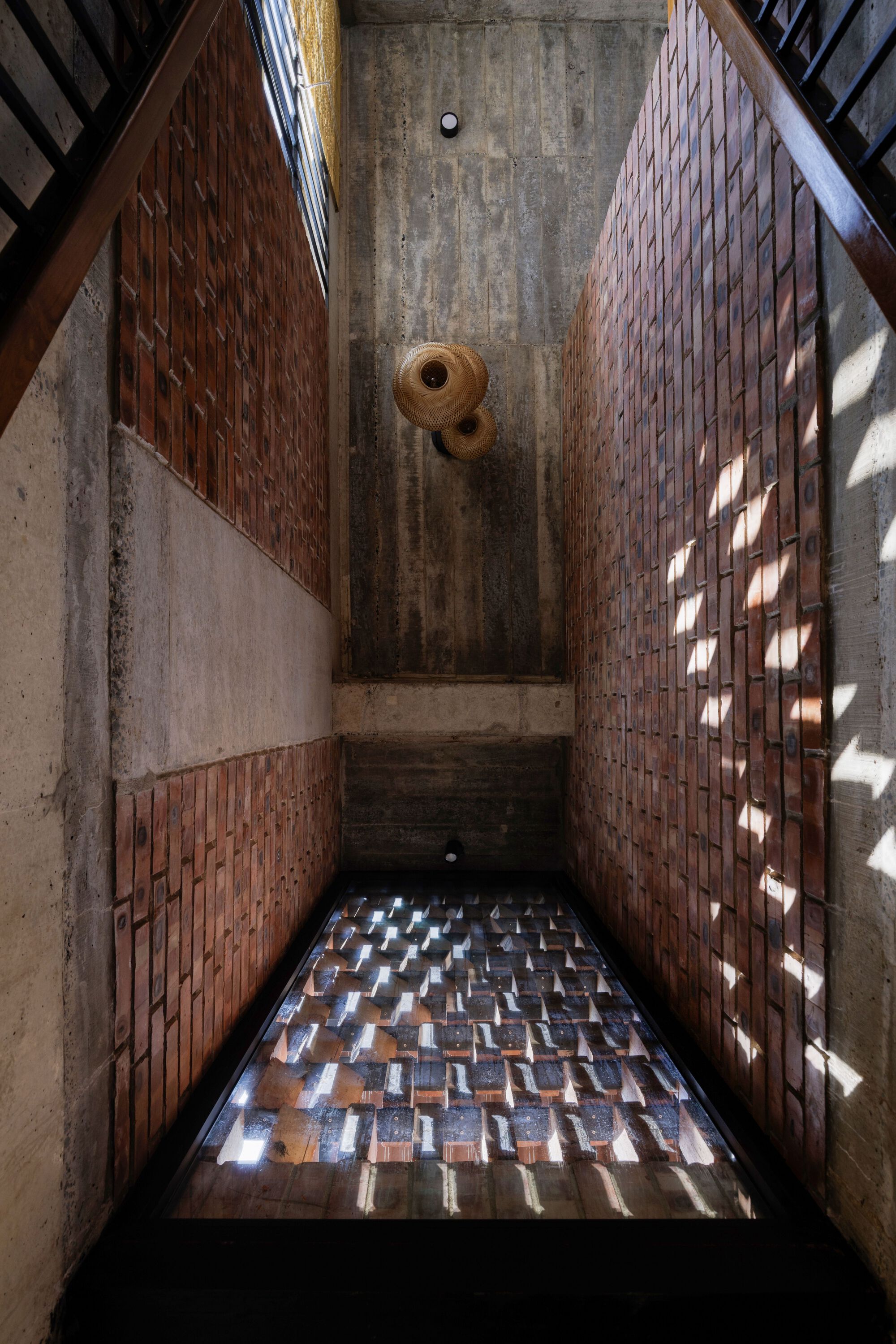 Looking down a tall concrete and brick stairwell where dappled sunlight falls on the brick wall and a glass floor panel
