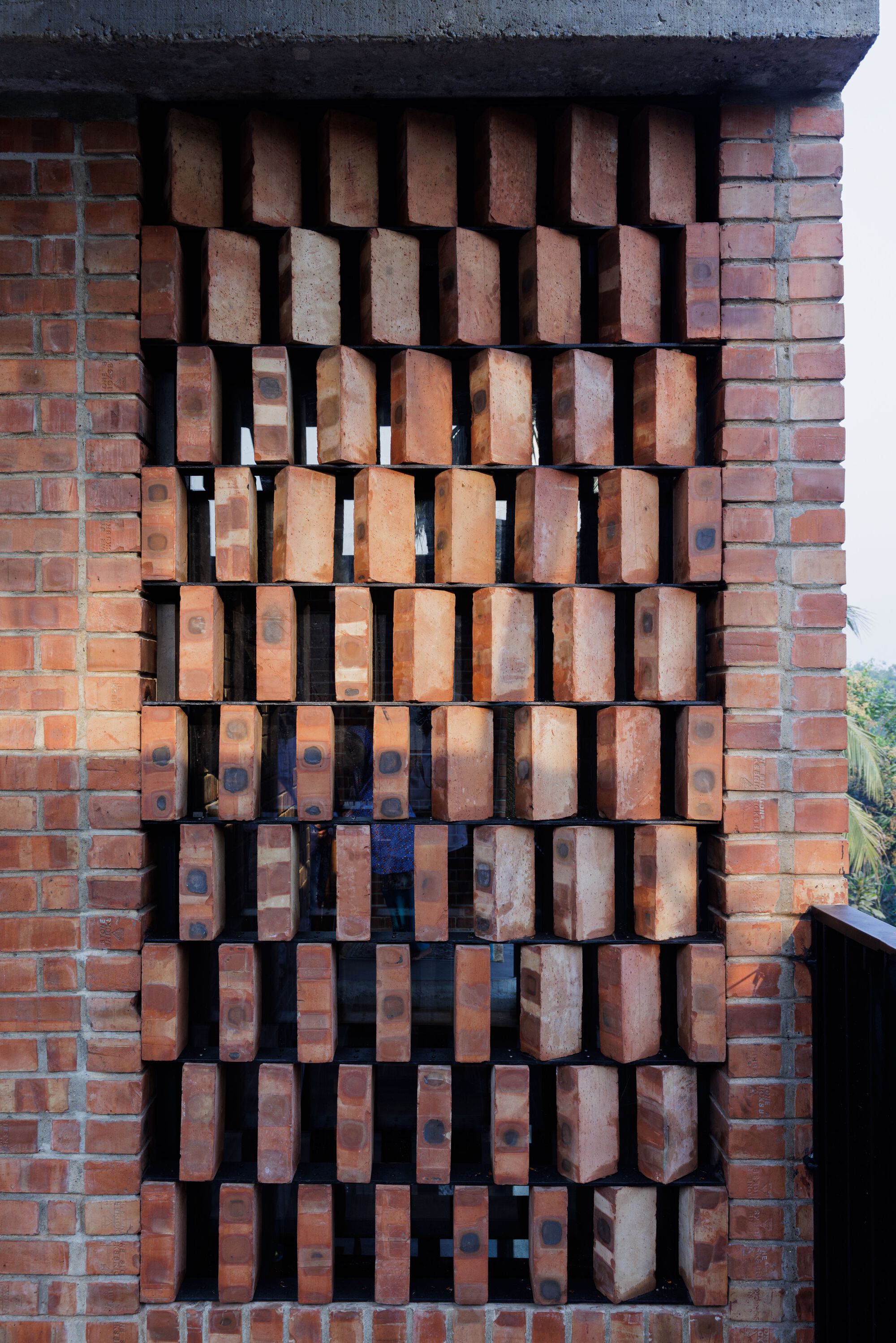 Close-up of a brick jali screen with rotated bricks set between solid brick piers, palm fronds visible beyond in daylight