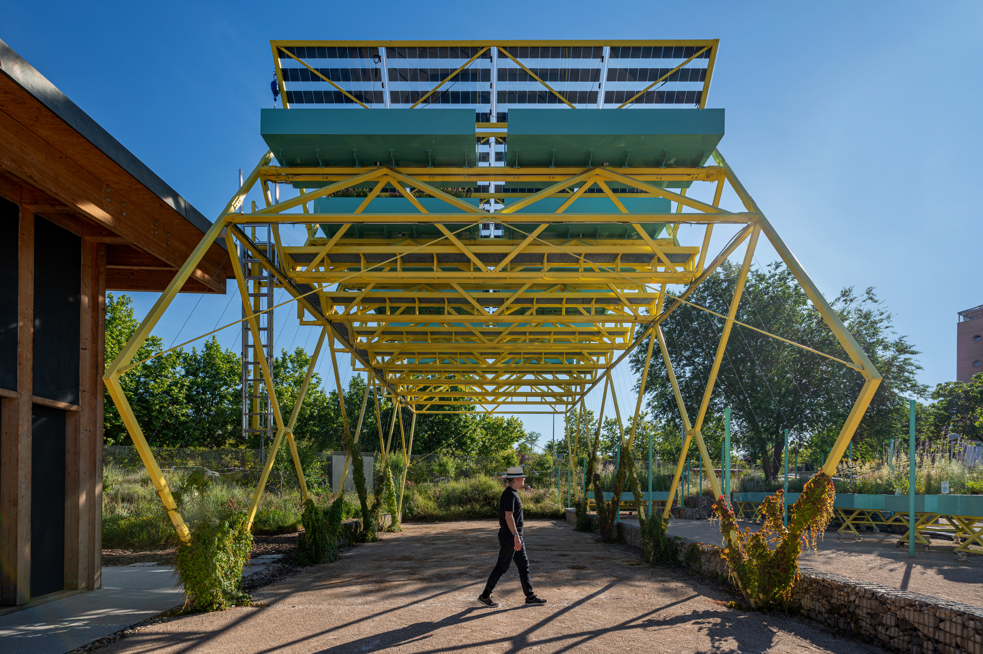 Yellow steel truss canopy carrying solar panels above a gravel path, with a walking figure and timber-clad building under a clear blue sky